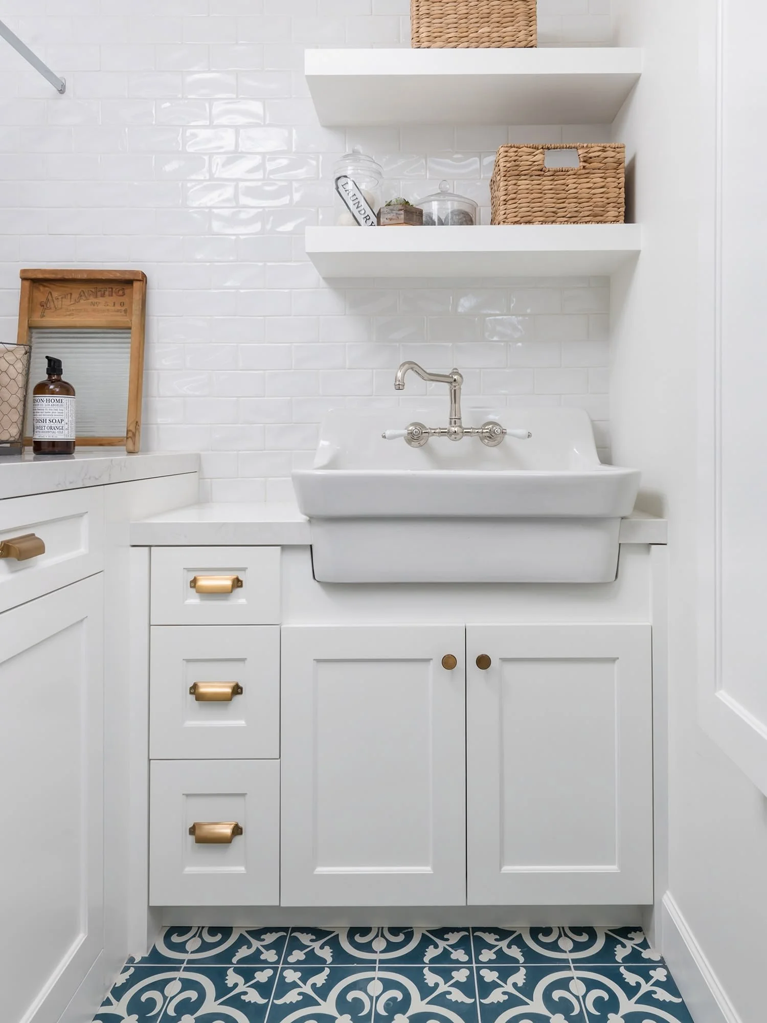 A white bathroom sink with gold handles on cabinets underneath. White tiled wall behind with floating shelves holding woven baskets and decorative items. Floor with black and white patterned tiles.