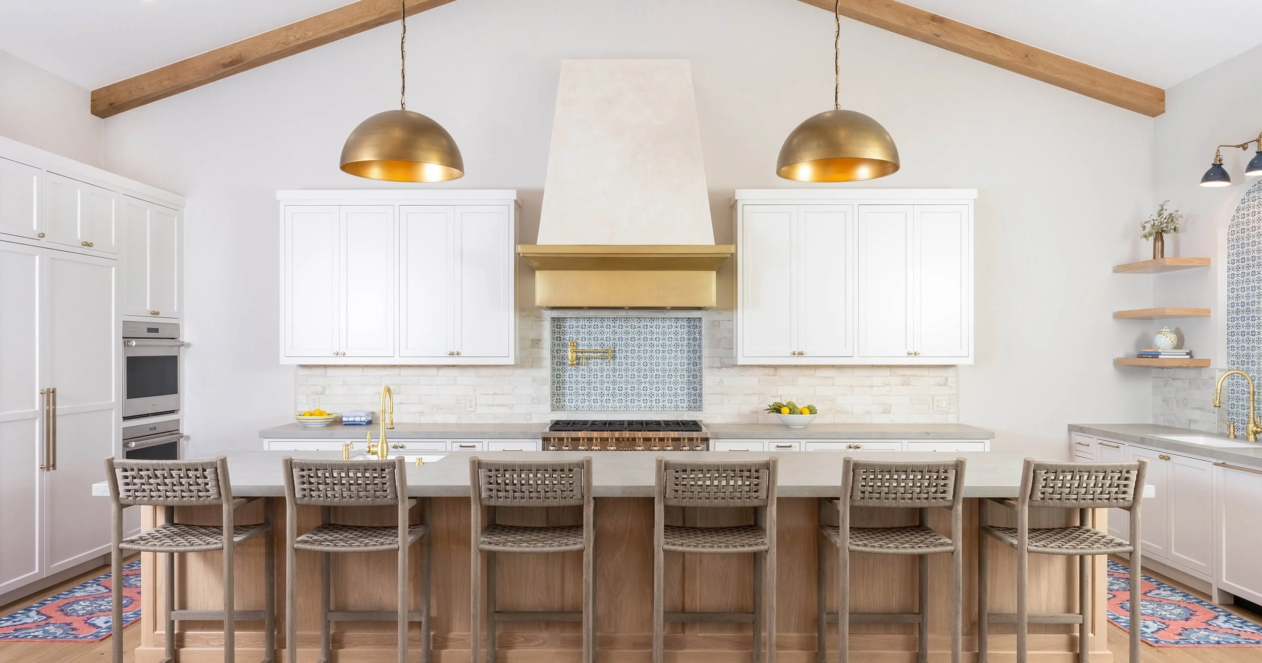 Modern white kitchen with a large island, gold accents, pendant lights, wood beams, and decorative shelves.