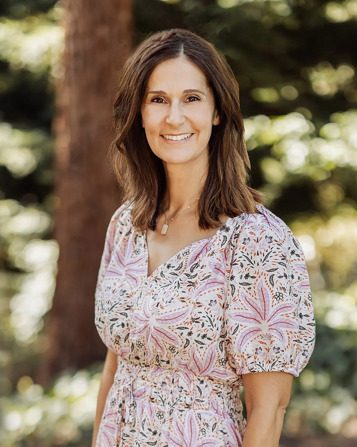 A woman with shoulder-length brown hair smiling outdoors, wearing a patterned blouse and a necklace, standing in front of a tree with blurred green foliage in the background.