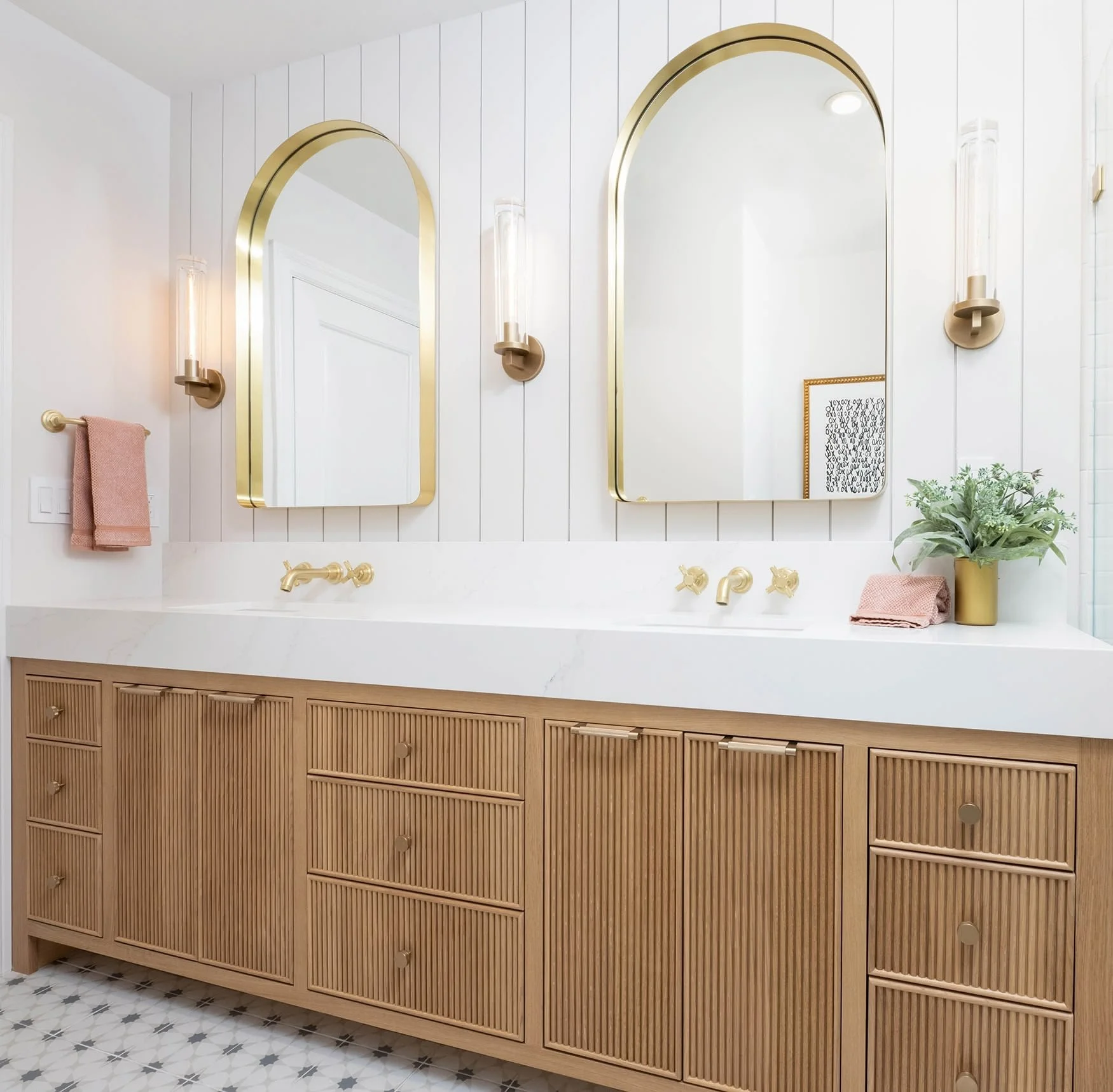Bathroom vanity with two large rectangular mirrors with gold frames, gold wall sconces, a beige rattan cabinet with multiple drawers, pink towels, a yellow vase with green plants, and a patterned tile floor.