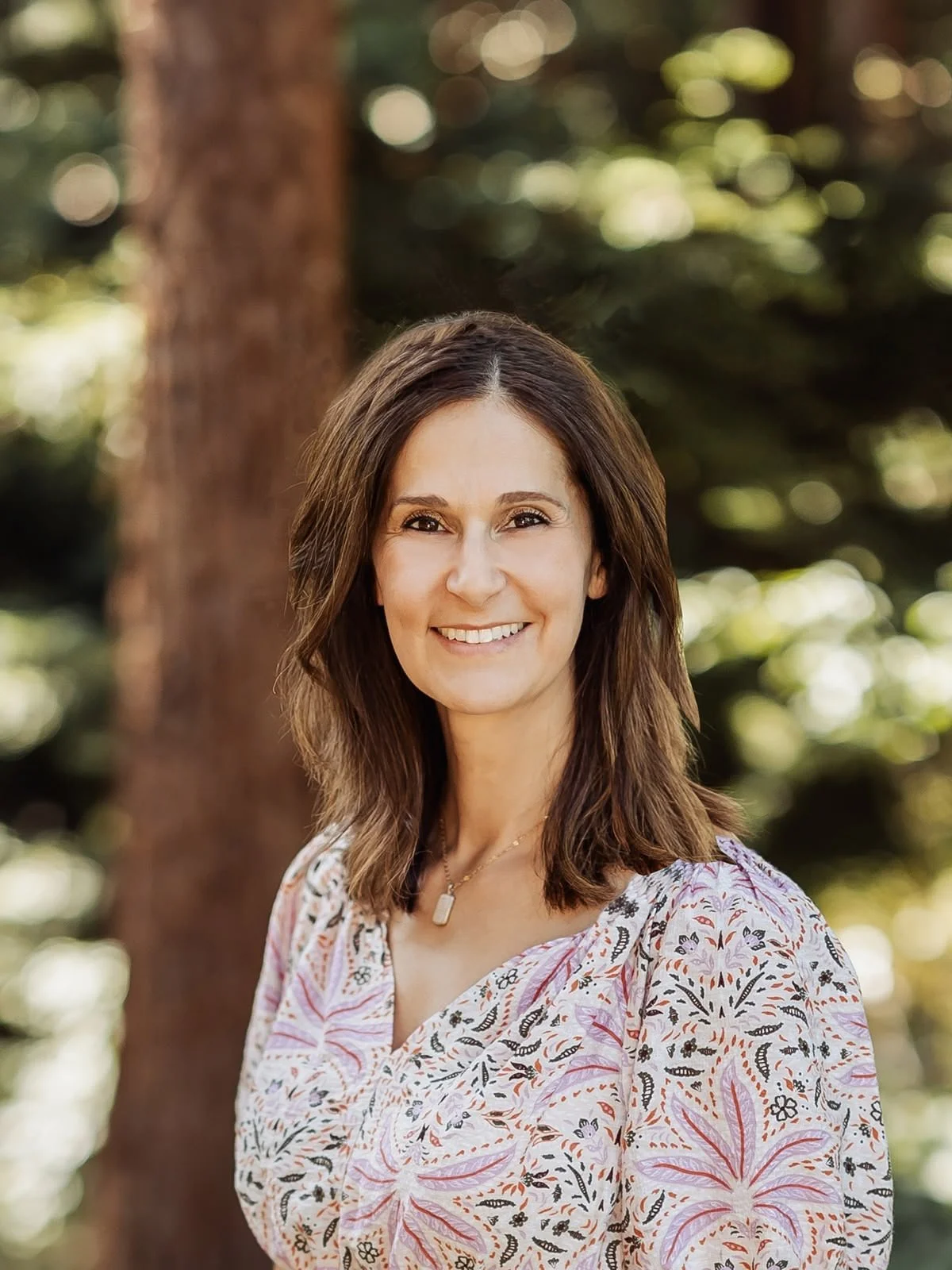 A woman with shoulder-length brown hair, smiling, outdoors with trees and green foliage in the background, wearing a patterned blouse and a necklace with a rectangular pendant.