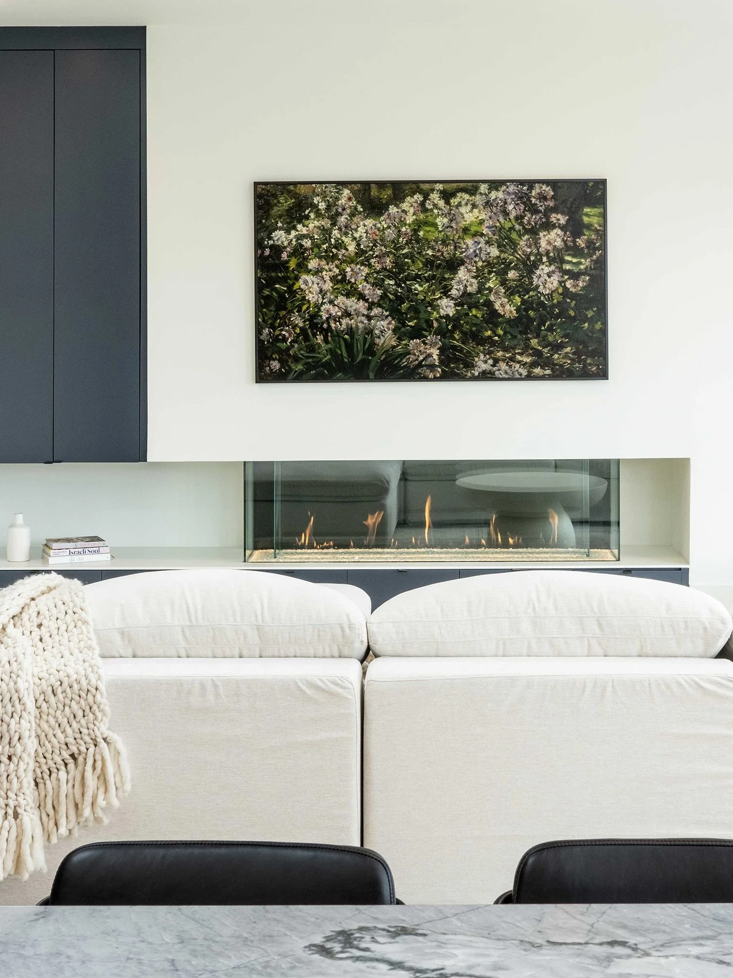 Living room with a white sofa, a knitted throw blanket, a fireplace, a framed floral picture, a bookshelf, and a marble table in the foreground.