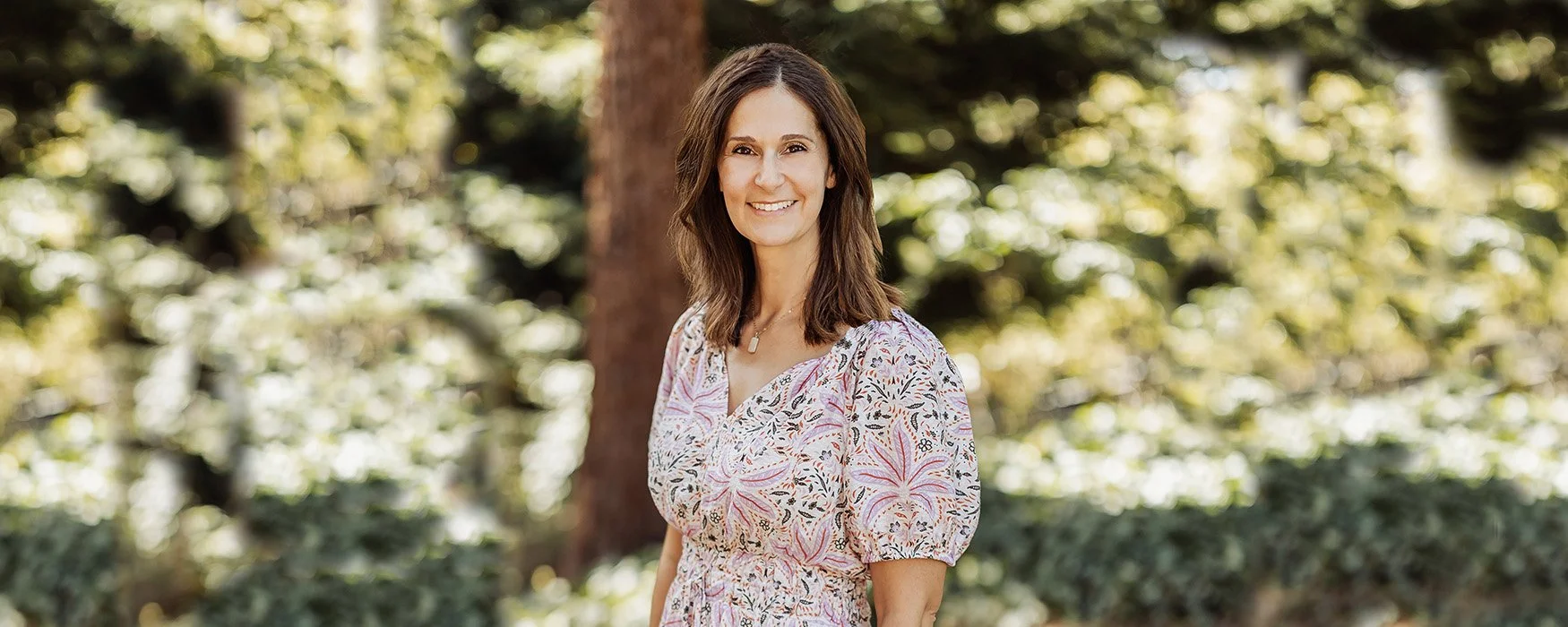 A woman with shoulder-length brown hair, smiling, standing outdoors in a park with blurred trees and greenery in the background.