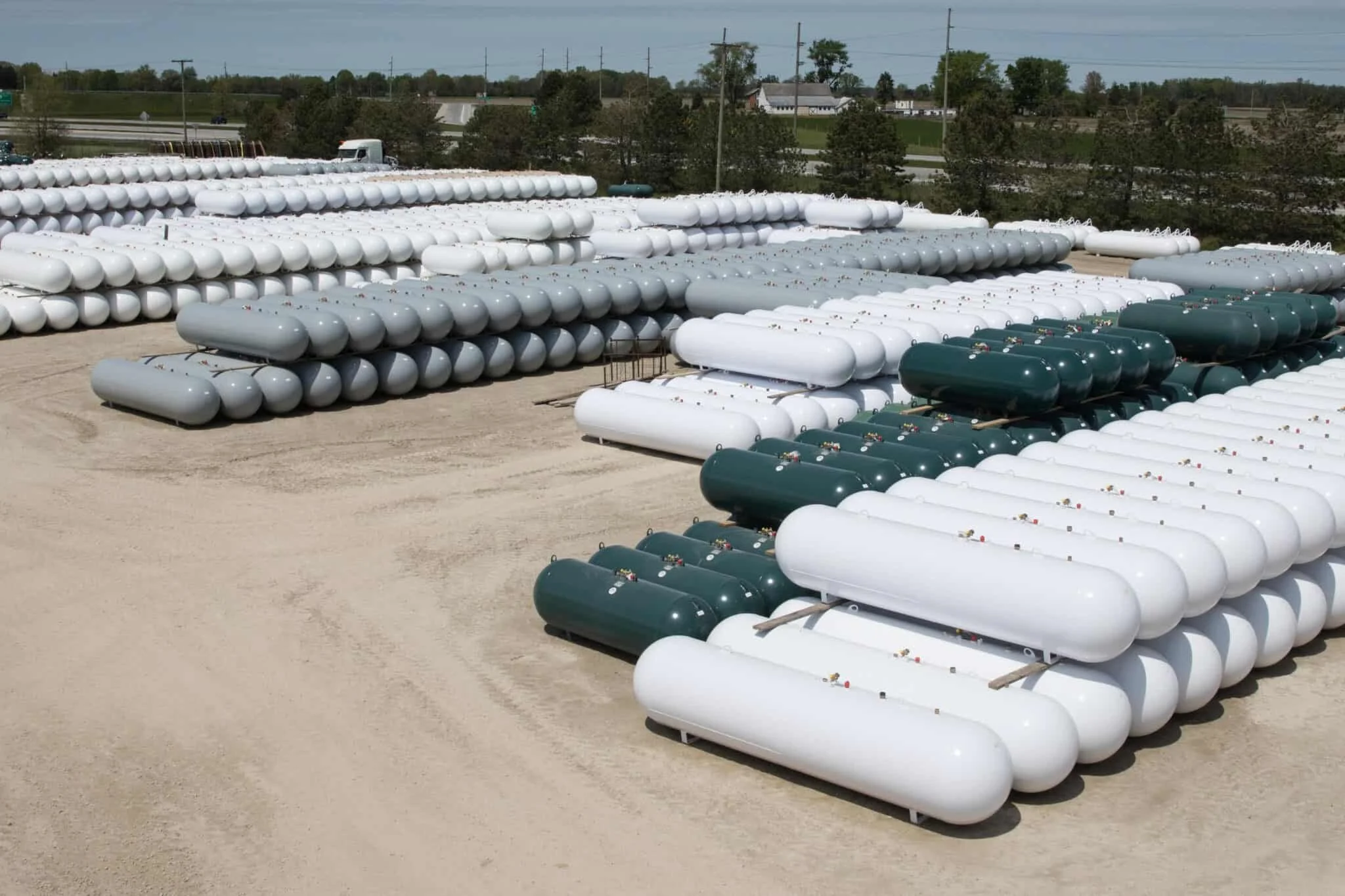 Multiple rows of white and green propane tanks are stored outdoors on a dirt lot, with a highway and trees in the background.