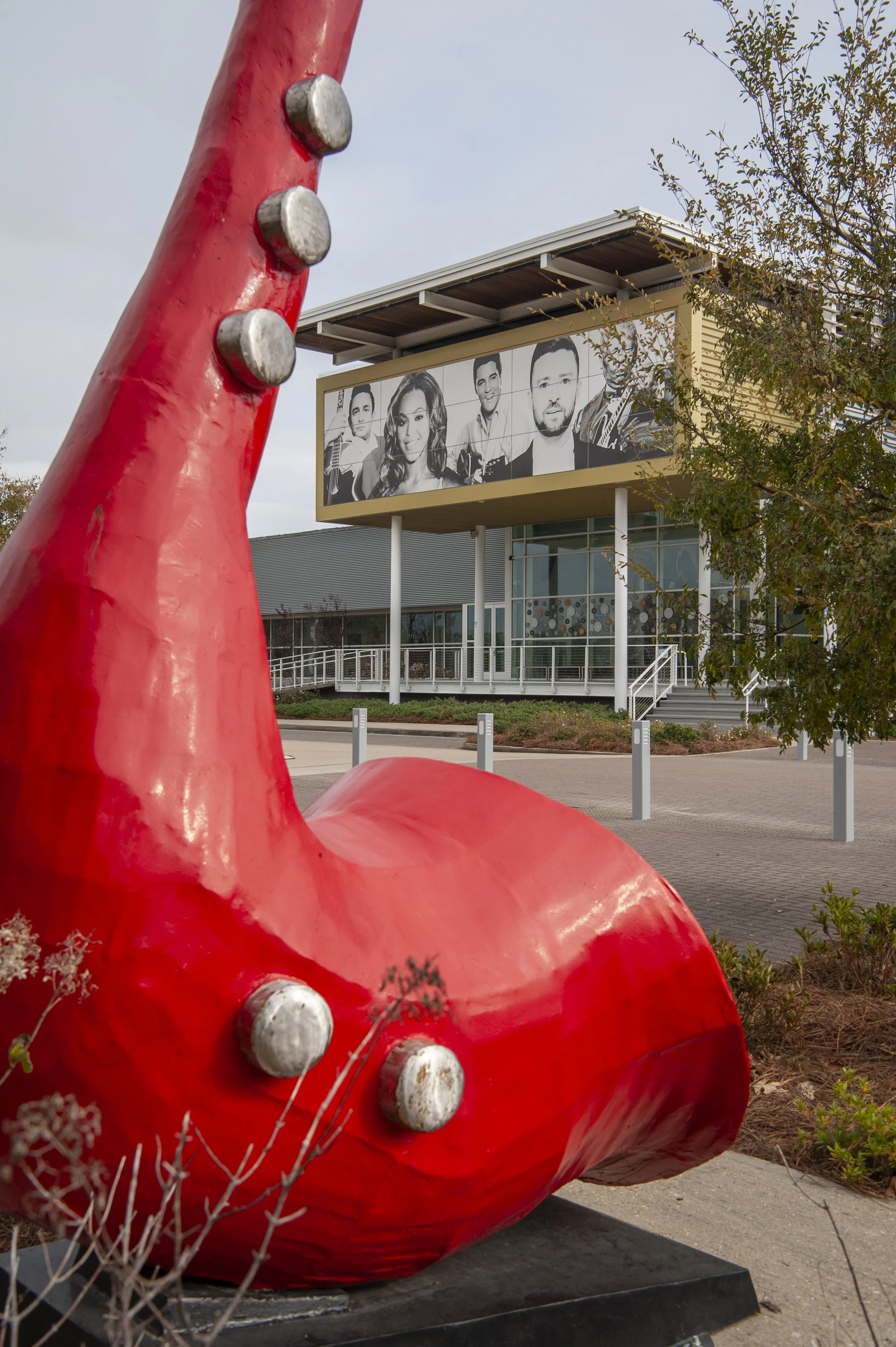 Close-up of a large red sculpture resembling a stylized animal, possibly a pig, with metal bolts on its body. In the background, there is a modern building with a large black and white billboard featuring images of diverse people, and some trees with leaves.