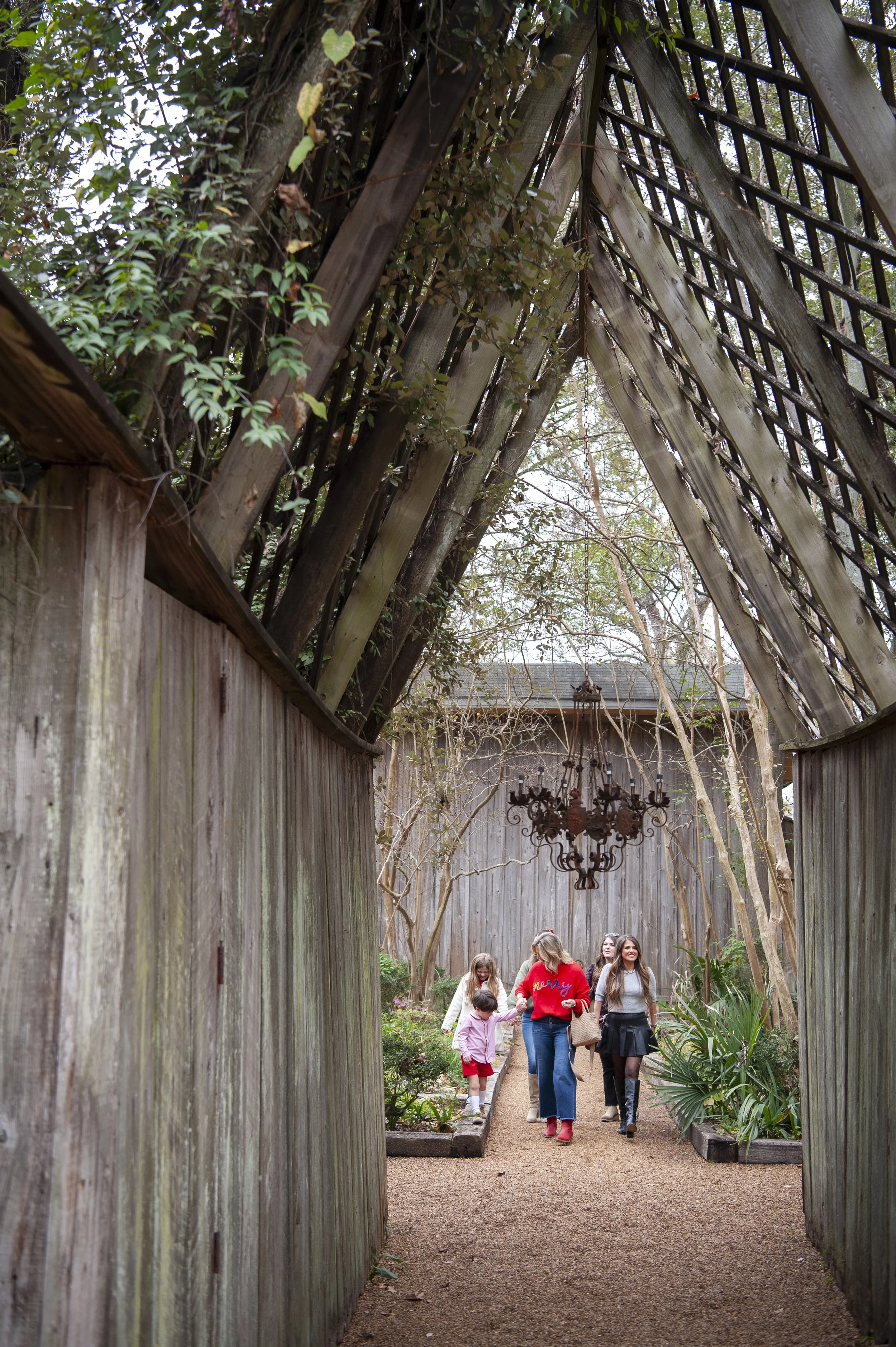 A group of children and adults walking through a narrow garden pathway enclosed by tall wooden fences with some greenery on both sides, and trees in the background.