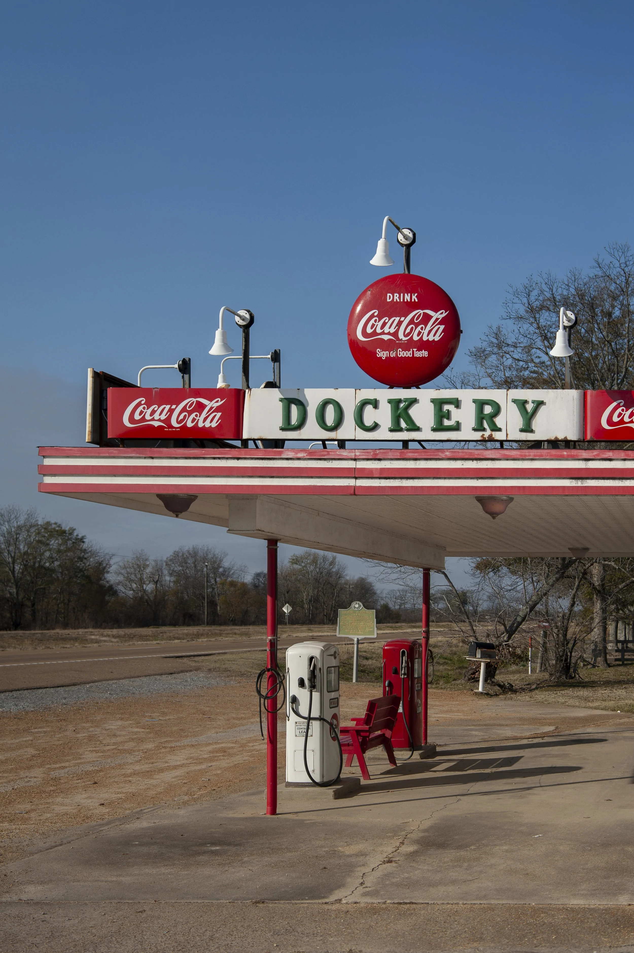 A vintage gas station with a large Coca-Cola sign on the roof and below it the store name 'Dockery'. There are multiple Coca-Cola signs, two gas pumps, a red chair, and small lamps on the roof. Tree in background, clear blue sky.