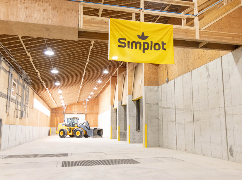 Empty industrial warehouse or storage facility with concrete walls, wooden ceiling, and yellow construction equipment in the background. A yellow banner with the Simplot logo hangs from the ceiling.
