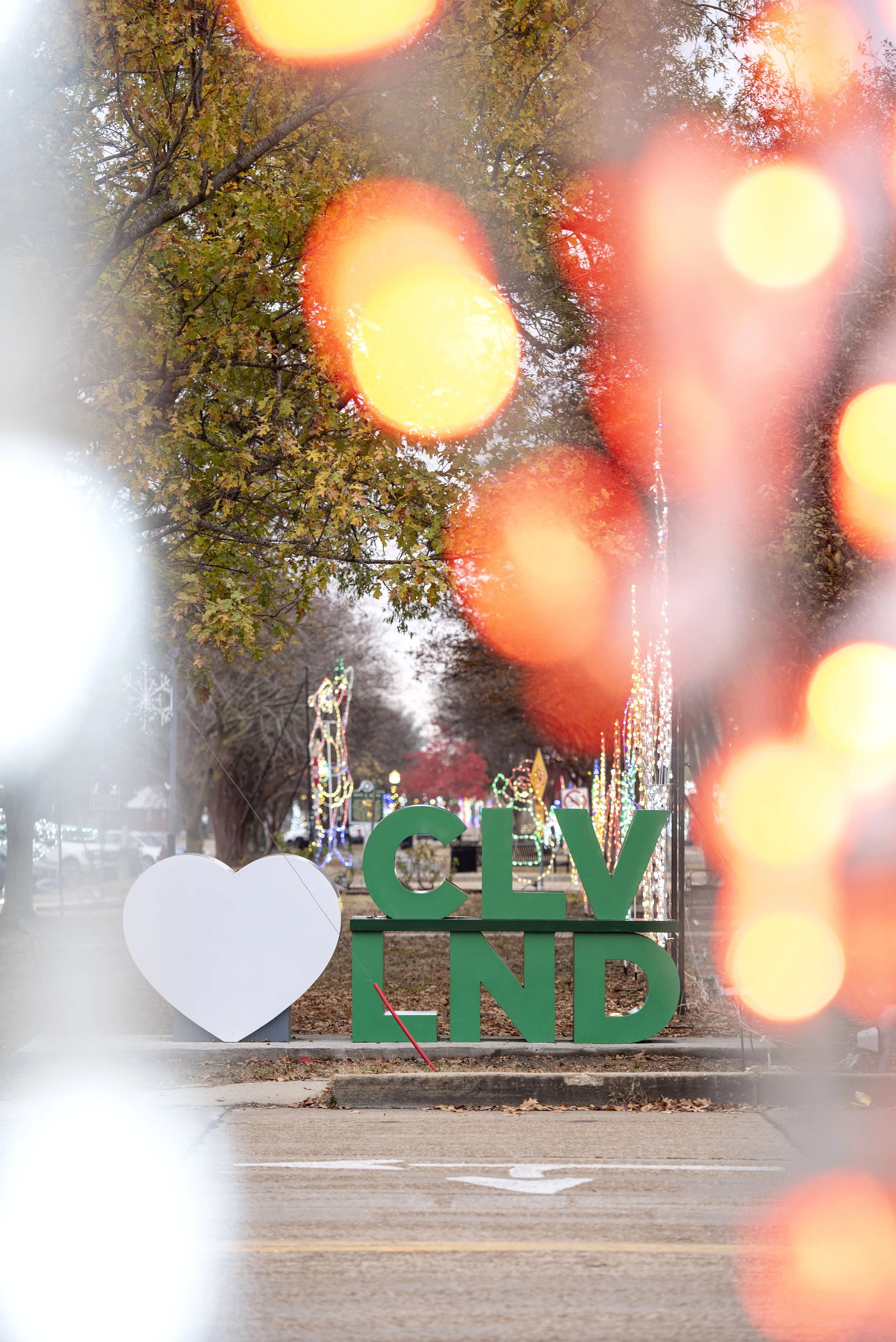 Decorative outdoor display with a white heart and green sign spelling 'CLV LAND', set against trees with colorful holiday lights and blurred warm light spots.