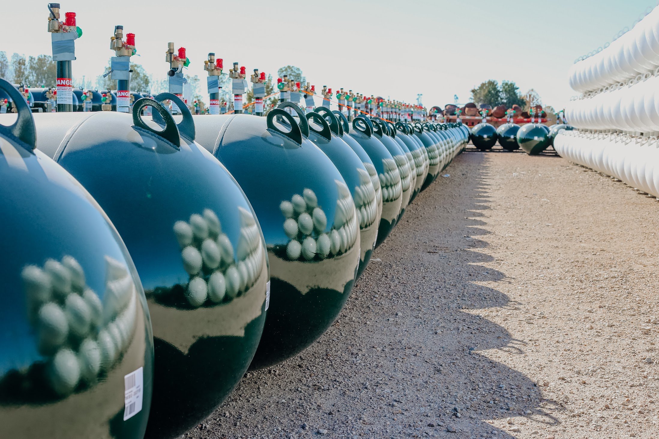 Large outdoor storage area with rows of large propane tanks, some with gauges and warning labels, and white coils of piping on the right.