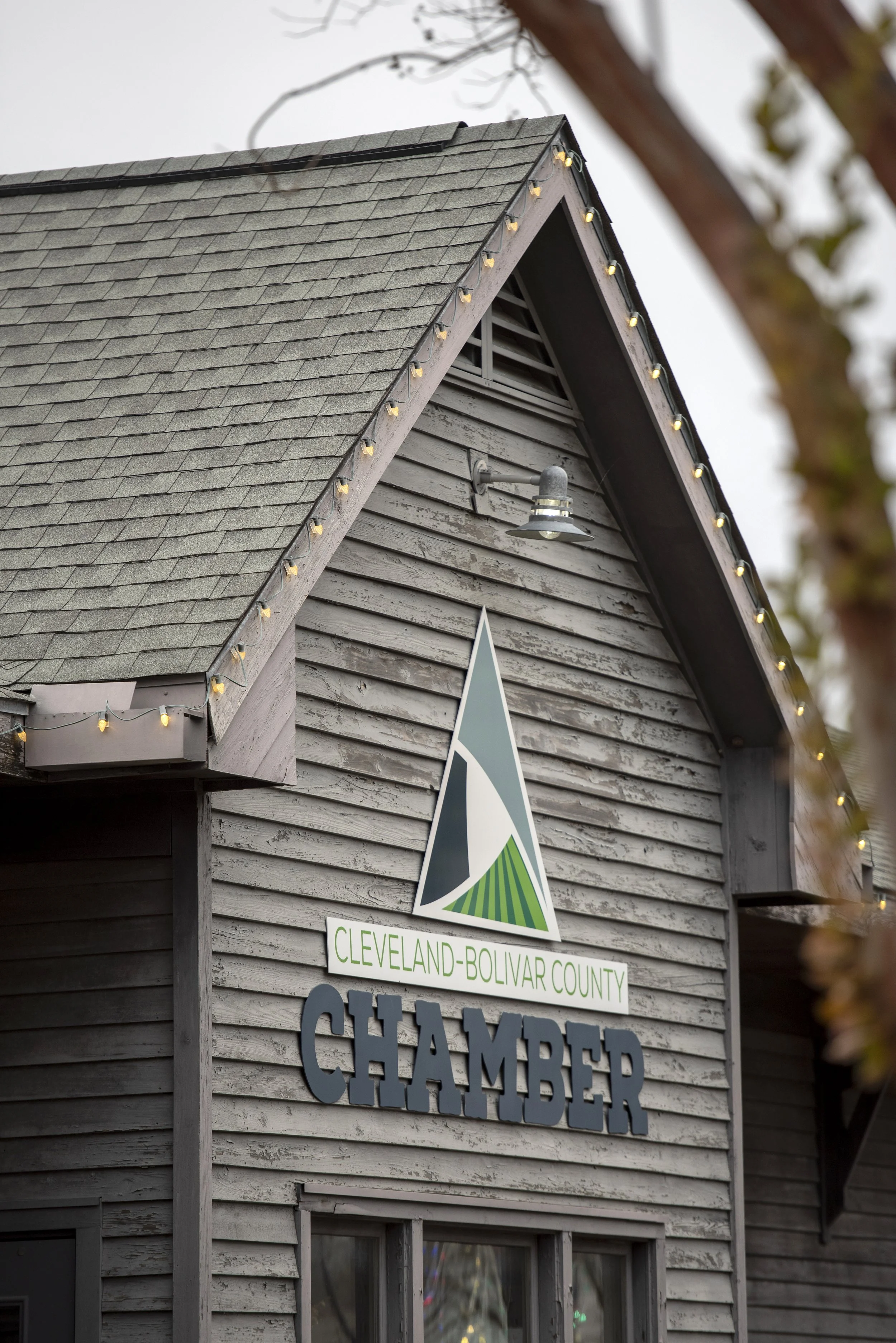 The exterior of a wooden building with a sign that reads "Cleveland-Bolivar County Chamber" and a logo with a mountain and green fields. The roof has string lights along the edge, and a vent and a light fixture are also visible on the building.