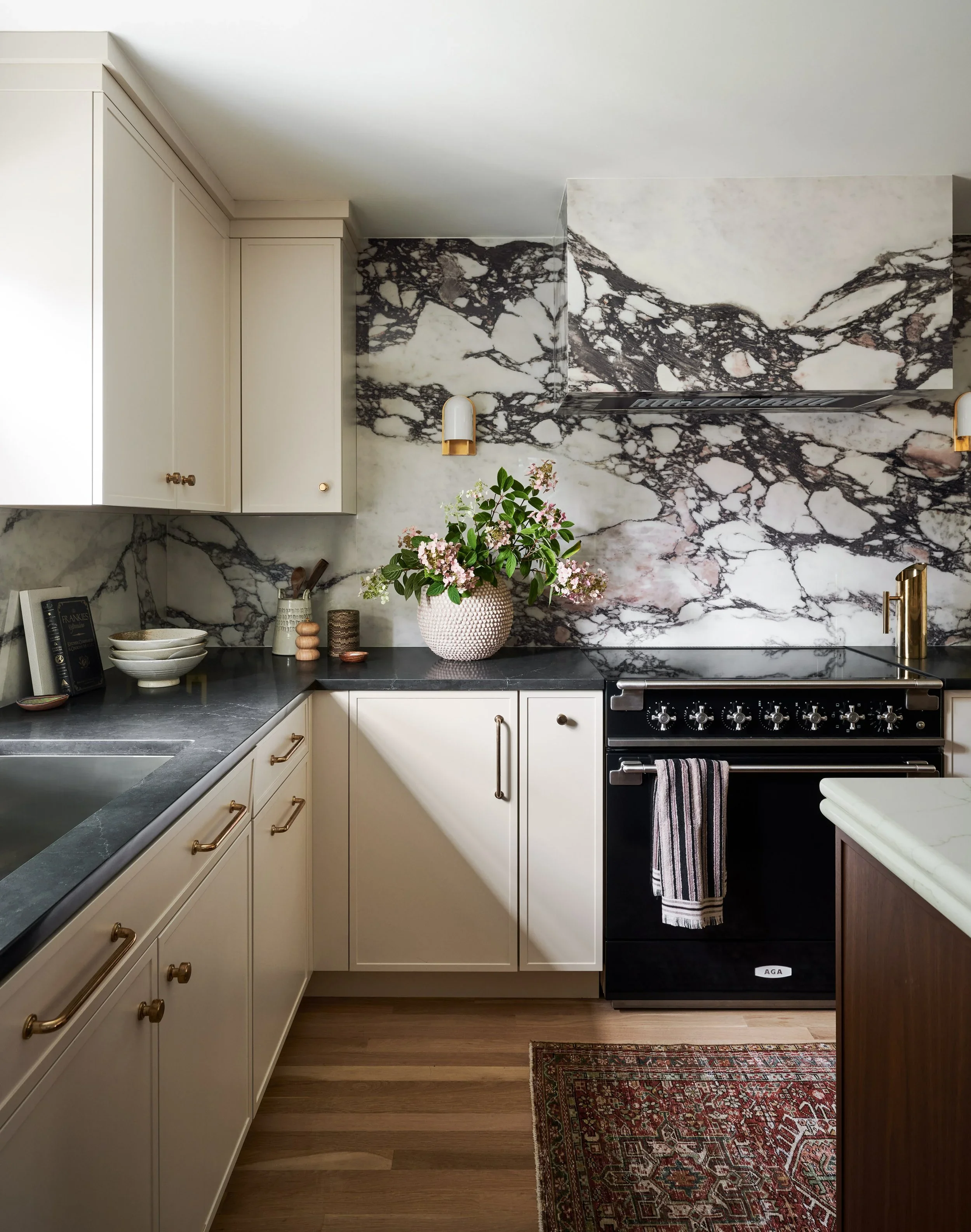 Kitchen with white cabinets, black countertops, a marble backsplash, a potted plant on the counter, and a black AGA oven, with a colorful patterned rug on the wooden floor.