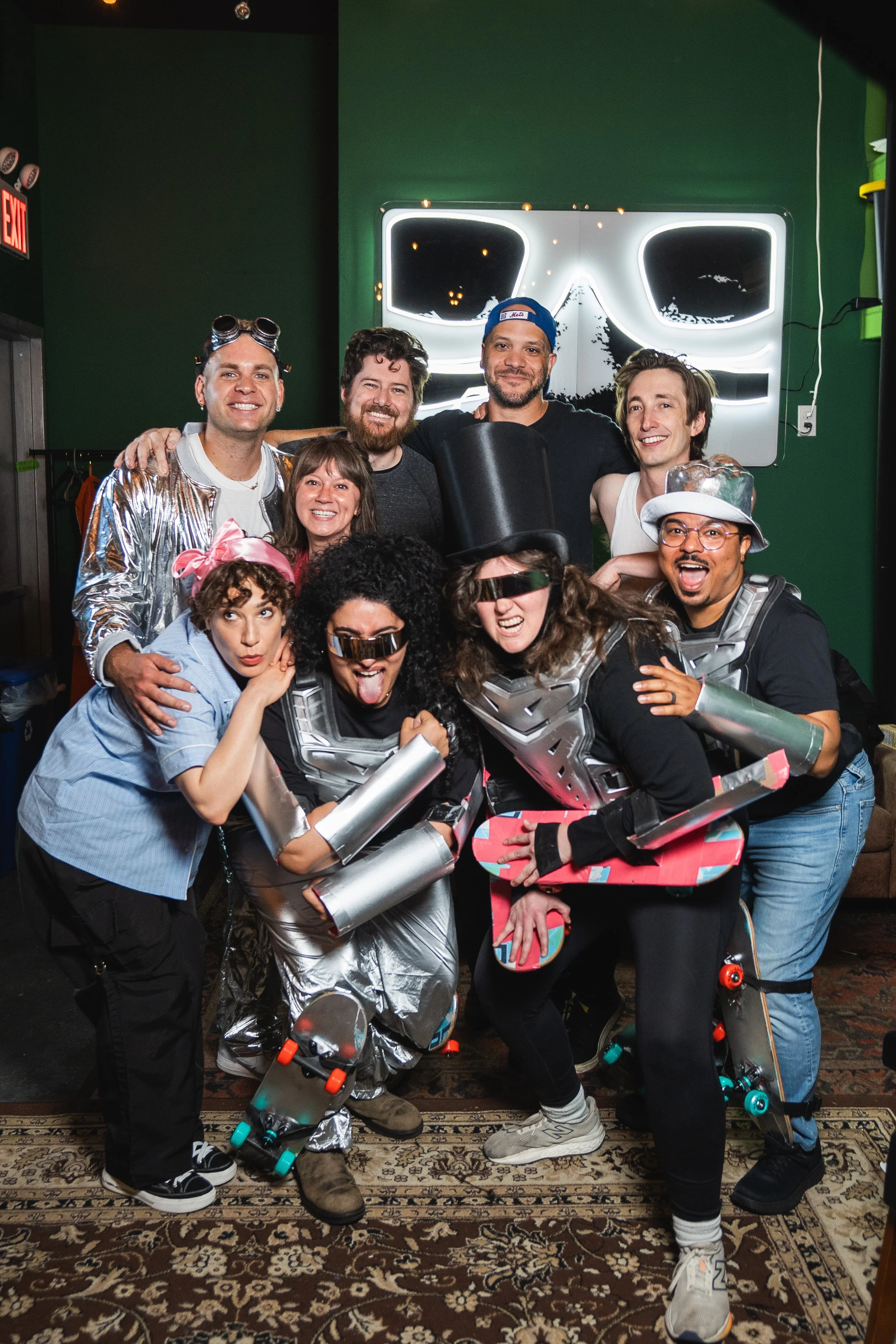 A group of nine people dressed in futuristic and retro costumes, some with roller skates, posing indoors in front of a neon sign that looks like a stylized face. They are smiling, making silly faces, and appear to be having fun at a themed event or celebration.