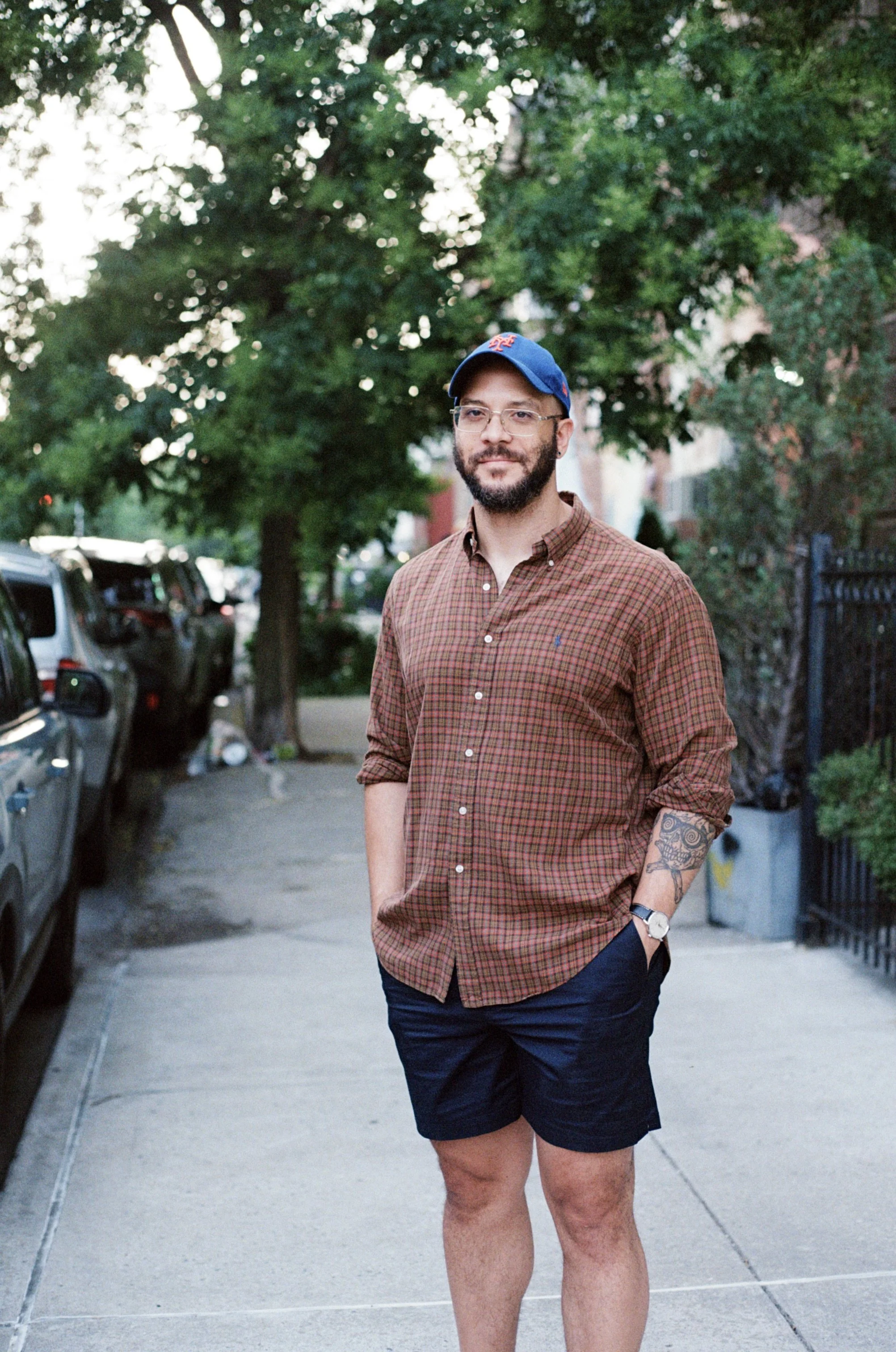 A man with glasses, a beard, and a mustache stands on a sidewalk with trees and parked cars behind him. He is wearing a blue baseball cap, a brown checkered button-up shirt, and navy shorts. His left arm has a tattoo, and he is smiling slightly while looking at the camera.