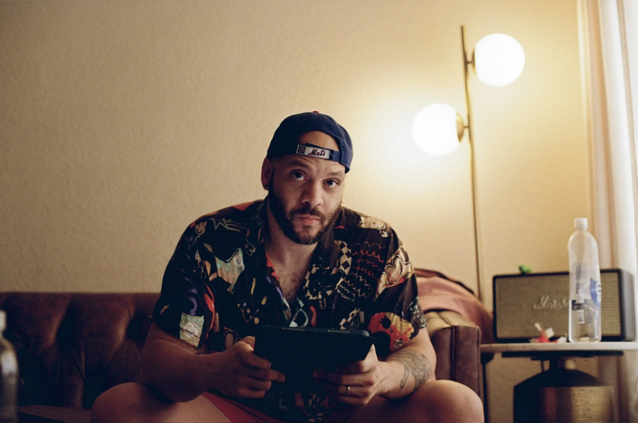 A young man with a beard wearing a black baseball cap backwards with 'Mets' logo, sitting on a brown leather couch in a dimly lit room, holding a tablet, wearing a colorful patterned short-sleeve shirt and pink shorts, looking at the camera. In the background, a floor lamp with three white globes, a window with curtains, a water bottle, a speaker, and some small items on a table.