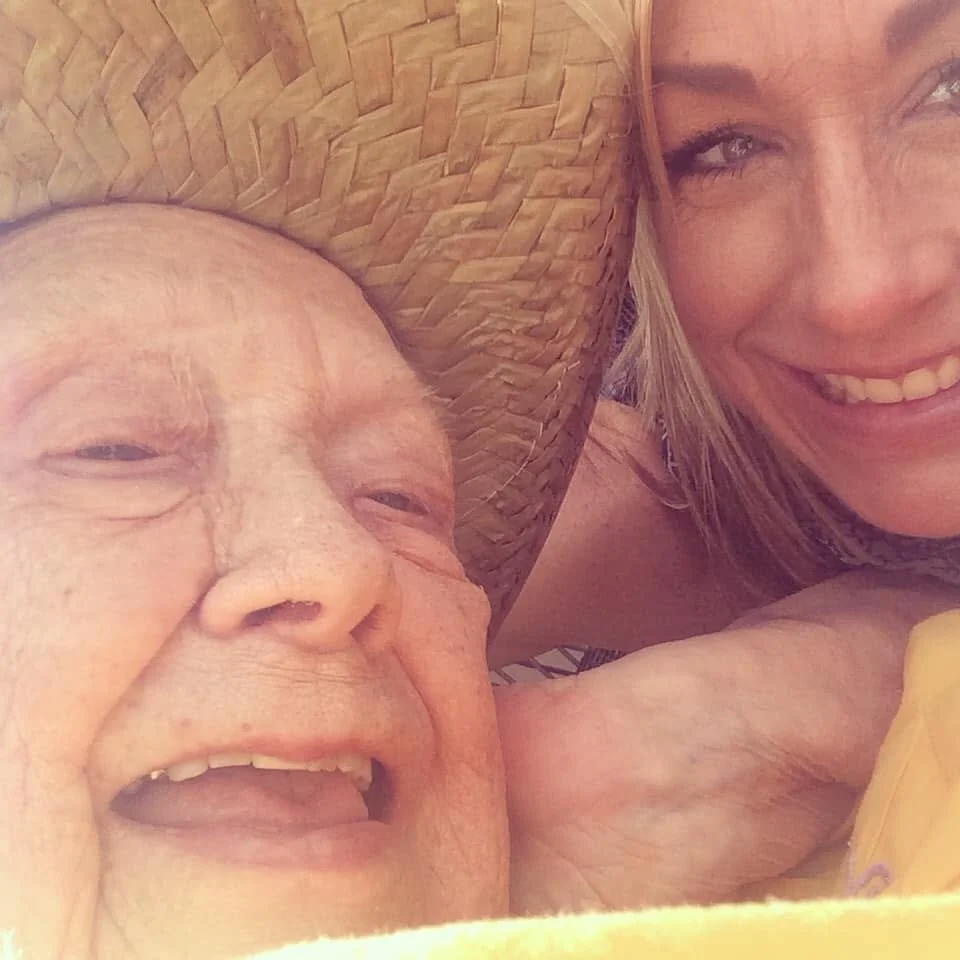 A close-up of an elderly woman and a younger woman lying down together, with the elderly woman wearing a large woven straw hat and smiling, and the younger woman smiling as well, showing some teeth.