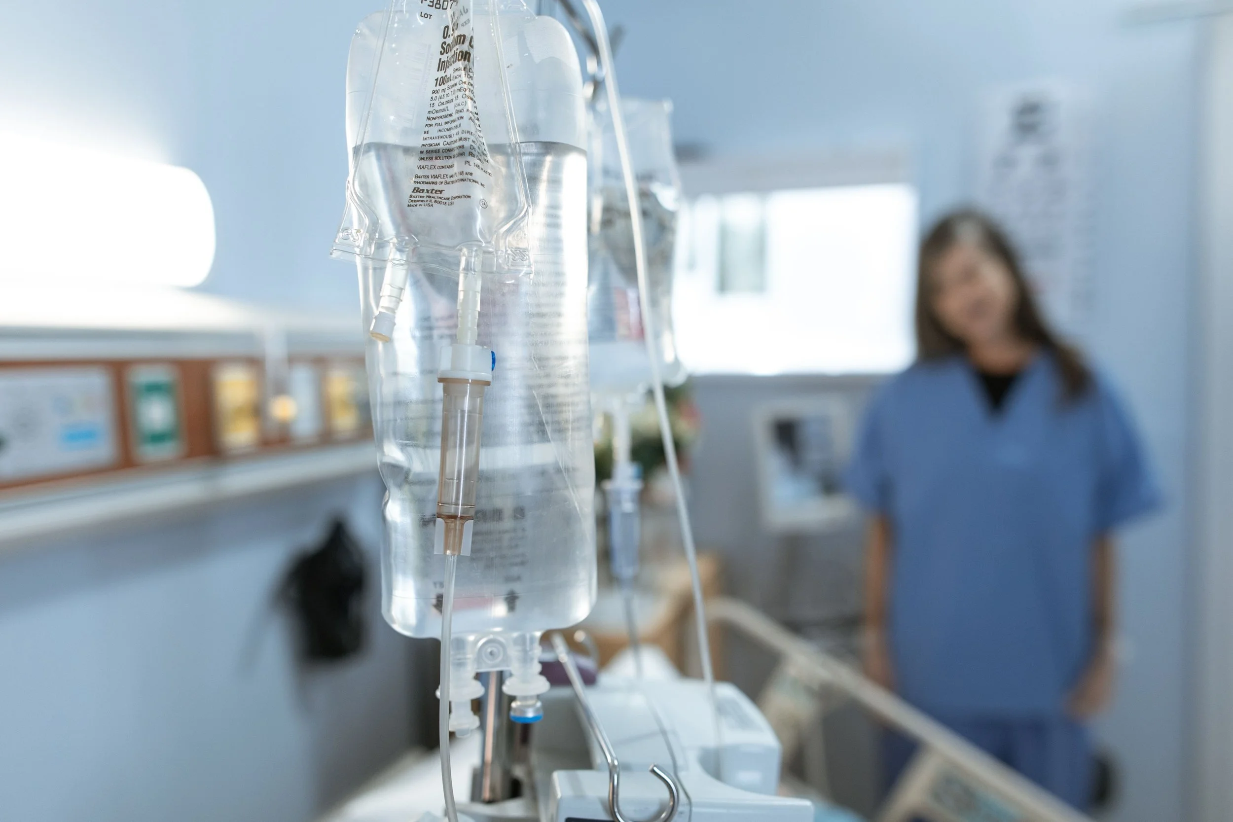 Close-up of an intravenous (IV) drip with a plastic bag and tubing hanging in a hospital setting, blurred background shows a woman in a hospital gown, smiling and talking to someone.