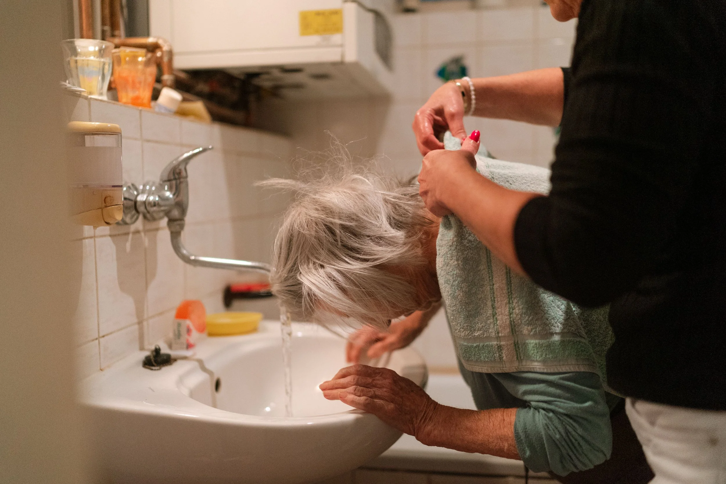 An elderly woman with gray hair washing her face at a bathroom sink while another person assists her, using a towel to help.