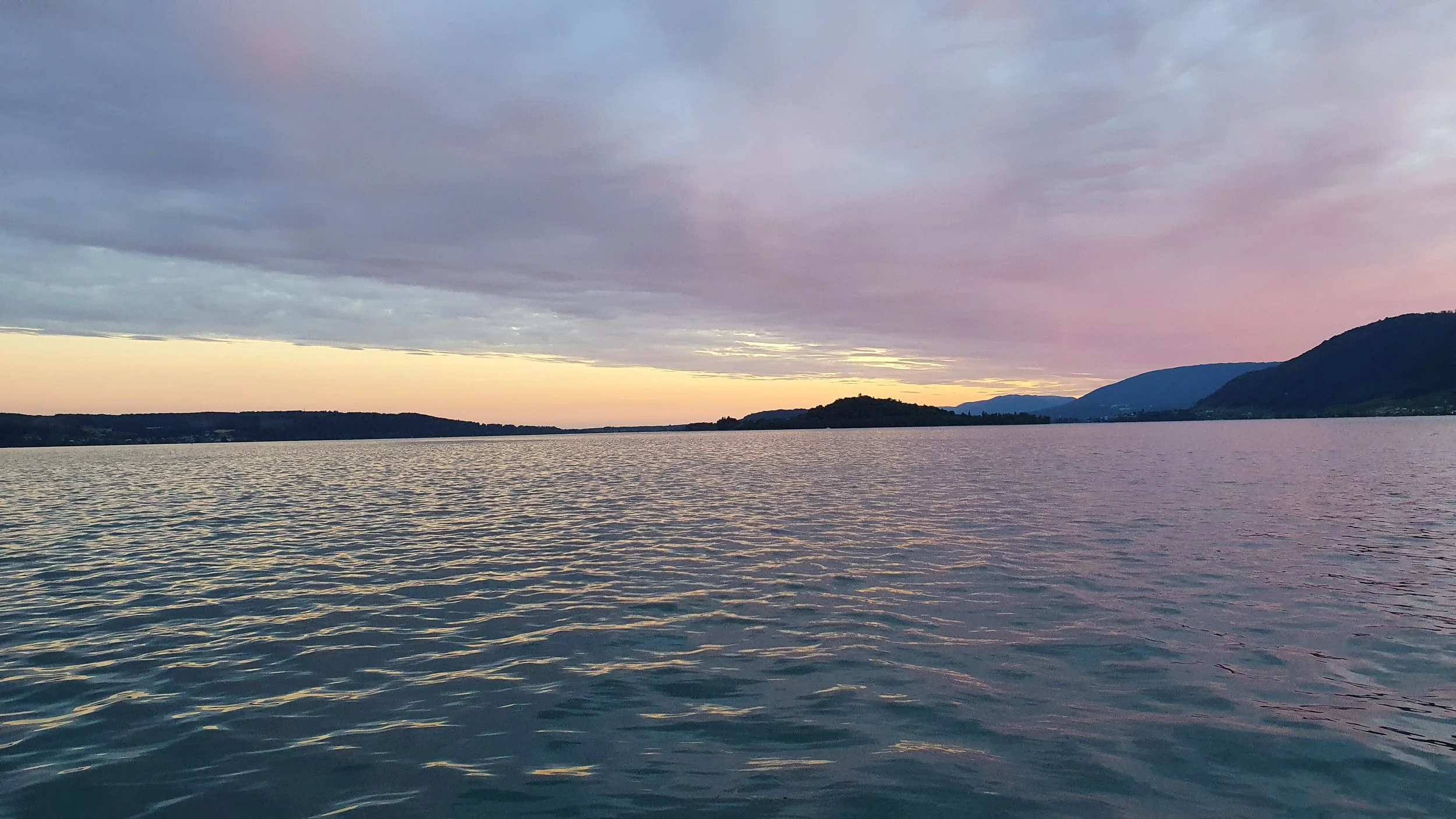 Ein ruhiger See bei Sonnenuntergang, mit Bergen und der St. Petersinsel im Hintergrund und einem bewölkten Himmel.