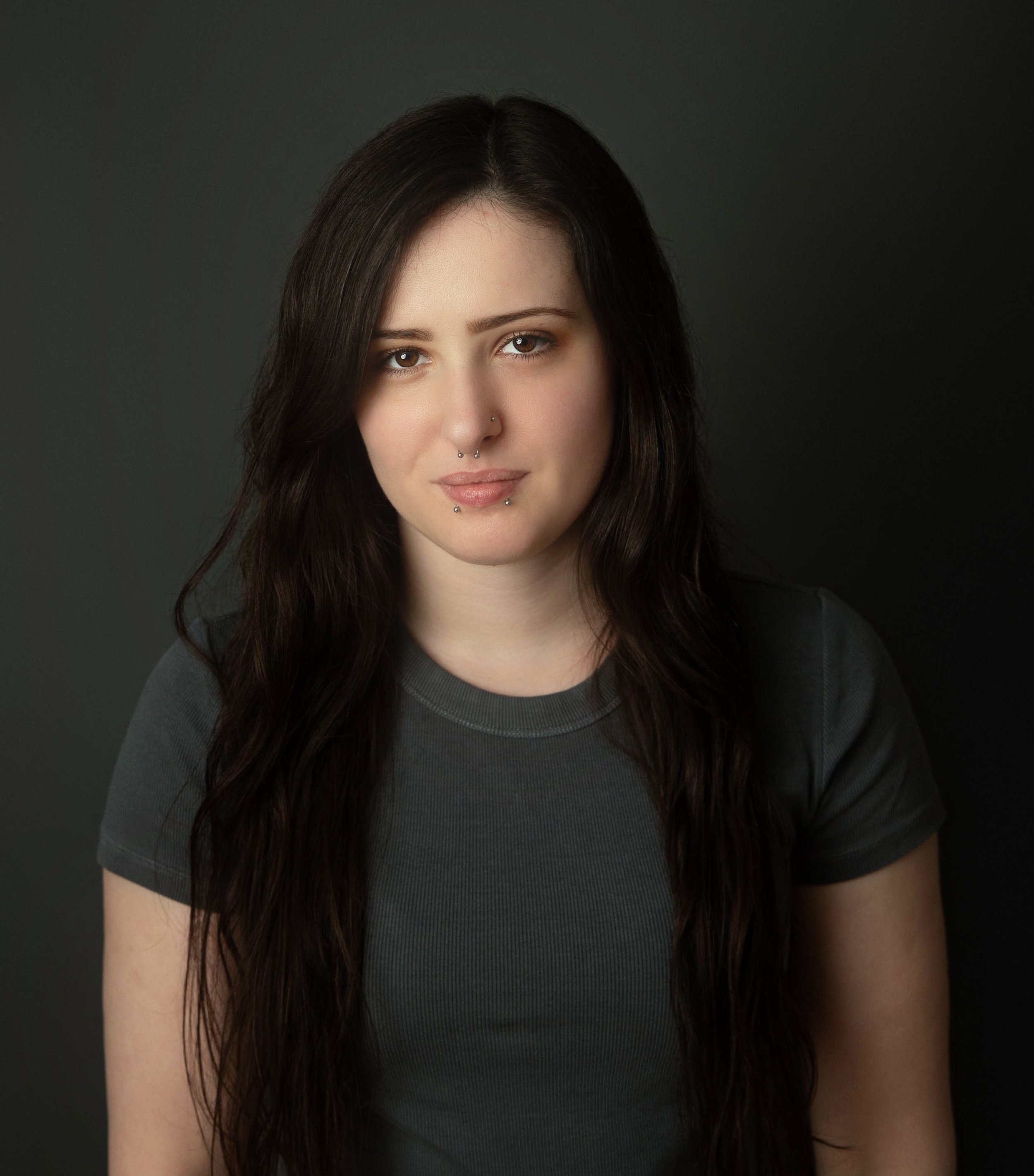 Head and shoulders portrait of a young woman with long dark hair, blue eyes, and several facial piercings, wearing a dark top, against a dark background.