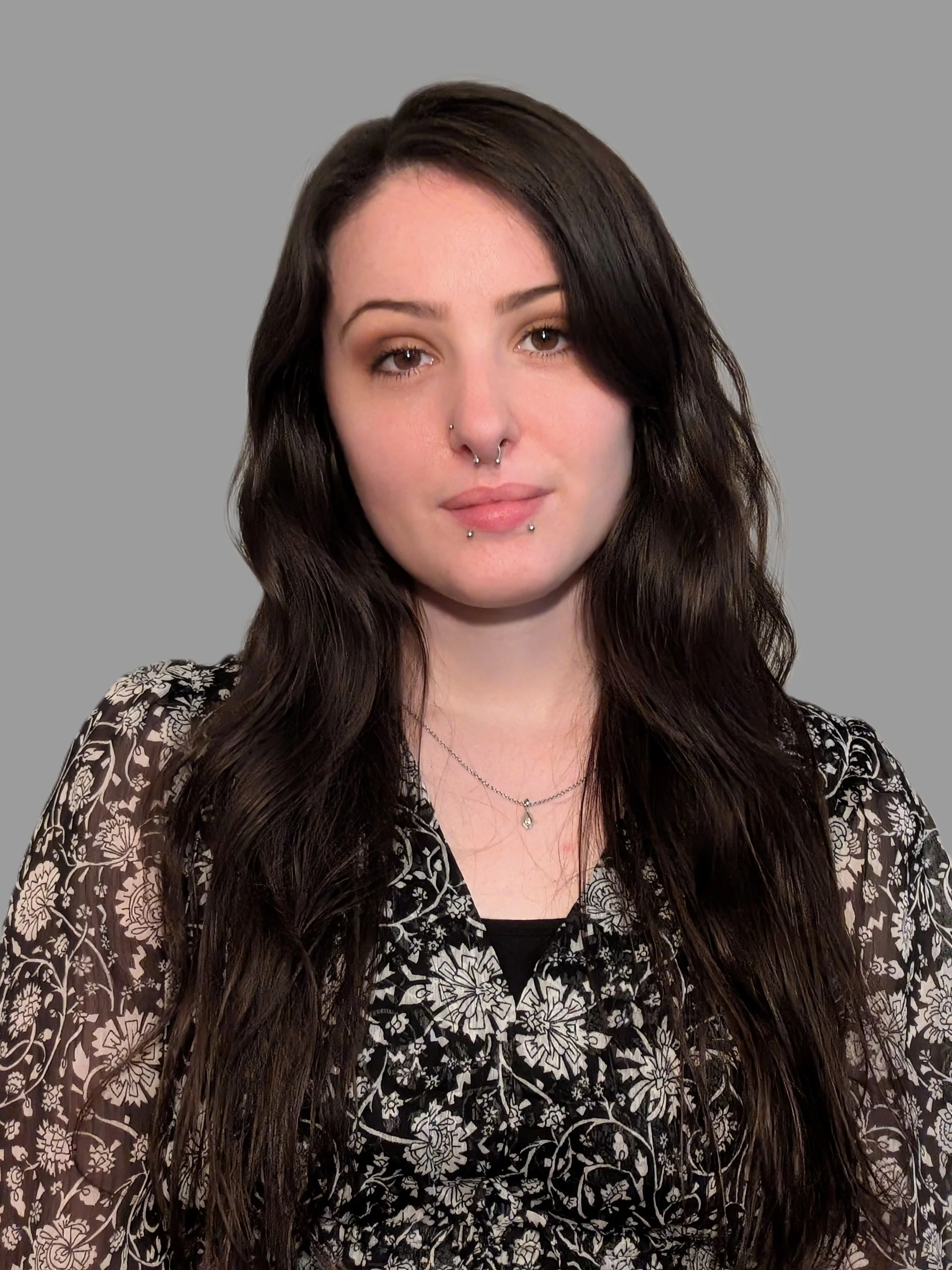 A woman with long dark hair, wearing a black and white patterned top, and wearing several facial piercings including a septum and labret, poses against a plain gray background.