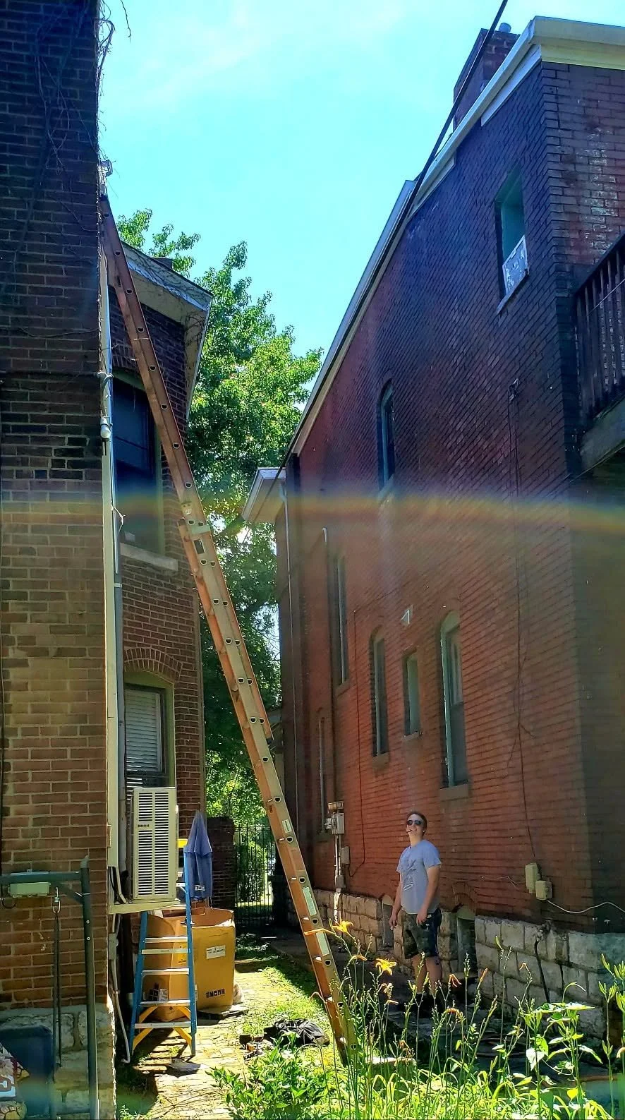 A man smiling and standing outside between two brick buildings with a ladder leaning against the left building. There is a rainbow glare across the photo from the sunlight.