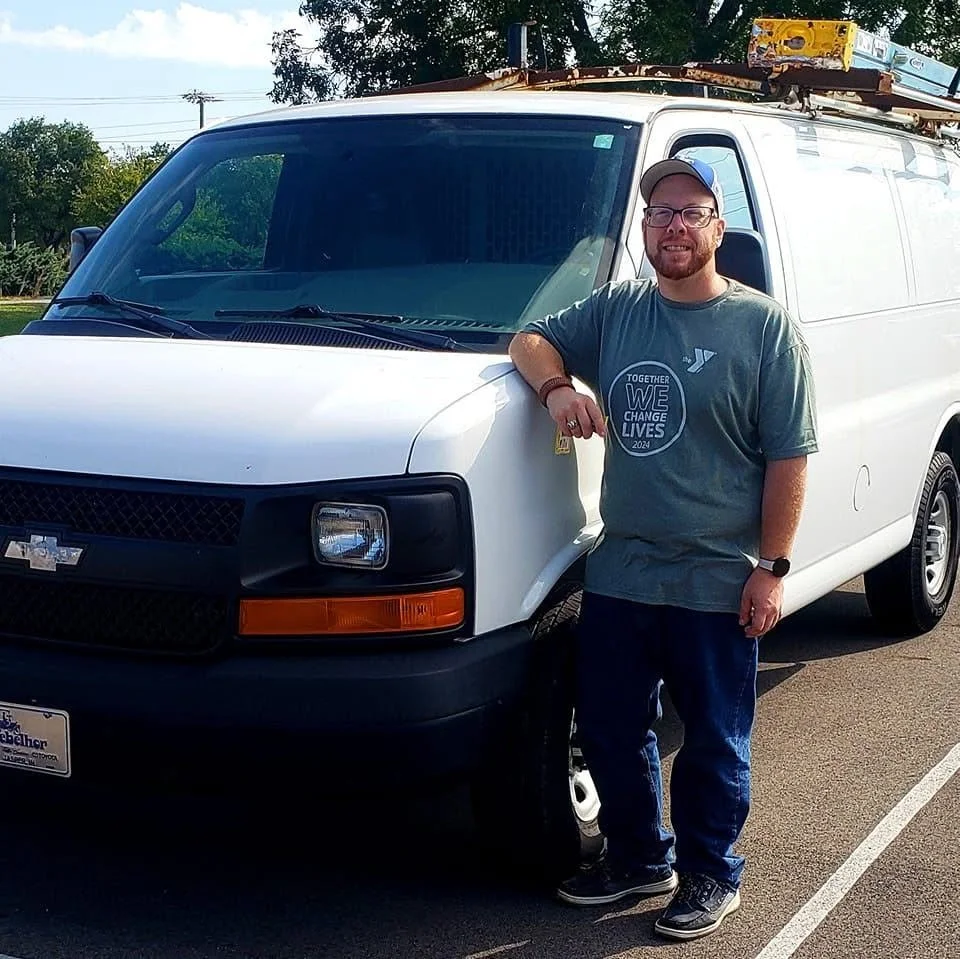 A smiling man with glasses, a cap, and a beard stands next to a white Chevrolet cargo van in a parking lot. He wears a gray T-shirt with the words 'TOGETHER WE CHANGE LIVES 2024' printed on it, blue jeans, and sneakers. The van has a ladder on the roof.