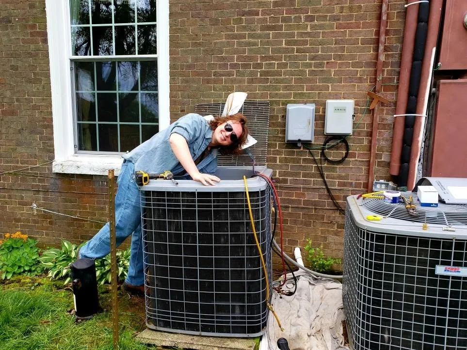 A woman wearing sunglasses and a blue shirt leaning on an outdoor air conditioning unit next to a brick house, smiling at the camera.