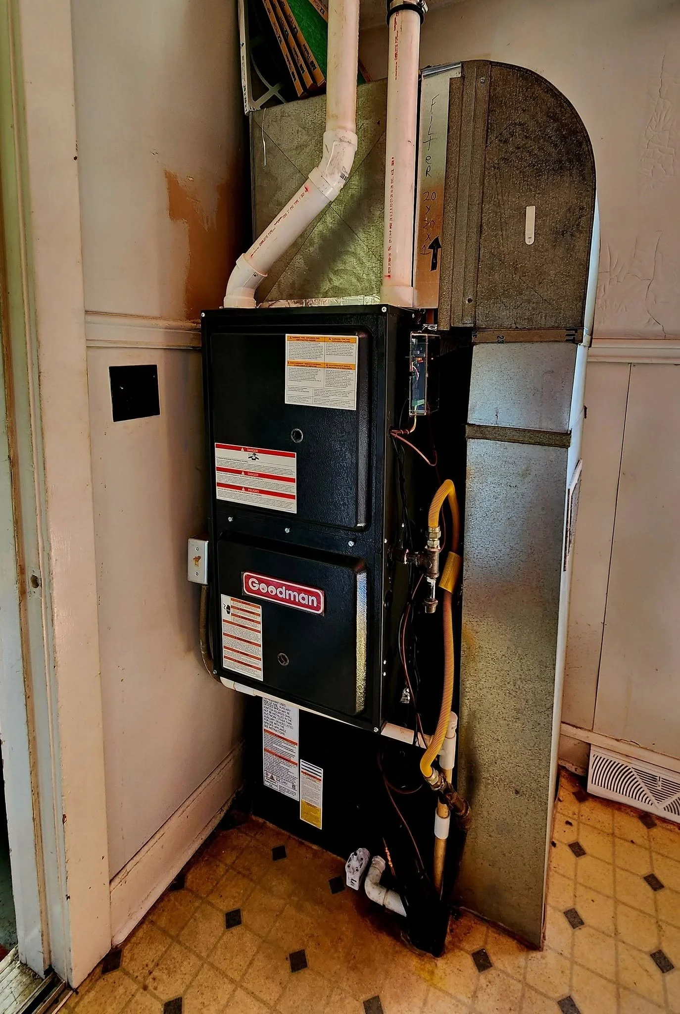 An HVAC furnace unit in a utility closet with pipes, ducts, and electrical wiring, situated on a tiled floor next to a wall and a door.