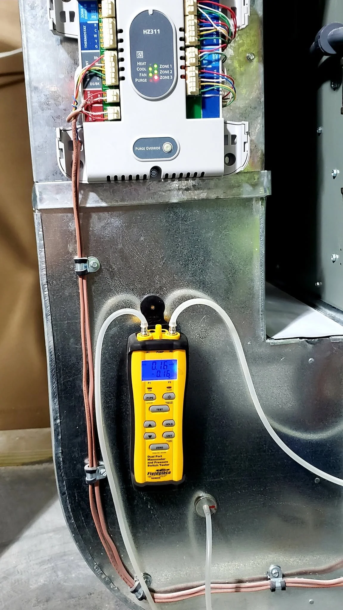 An HVAC control panel with wiring, a refrigerant gauge, and a digital manometer placed on a metallic surface.