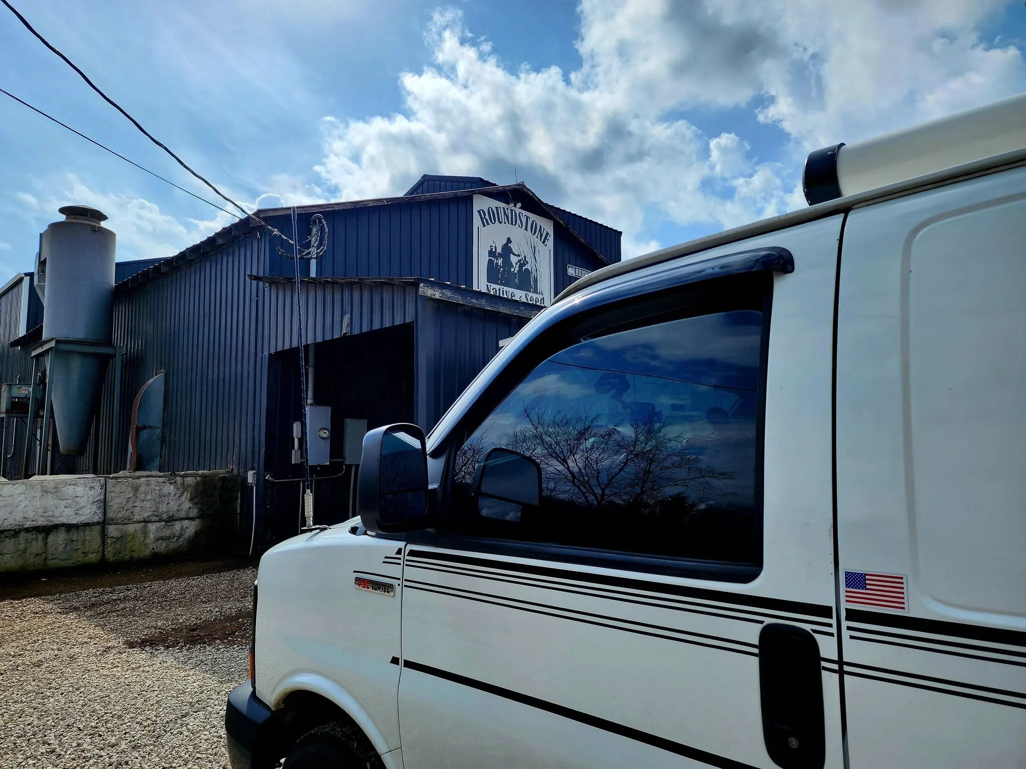 A white RV with black stripes and an American flag decal parked in front of a blue metal building with a sign that reads "Roundstone Native Seed." The sky is partly cloudy with blue patches, and power lines are visible above.