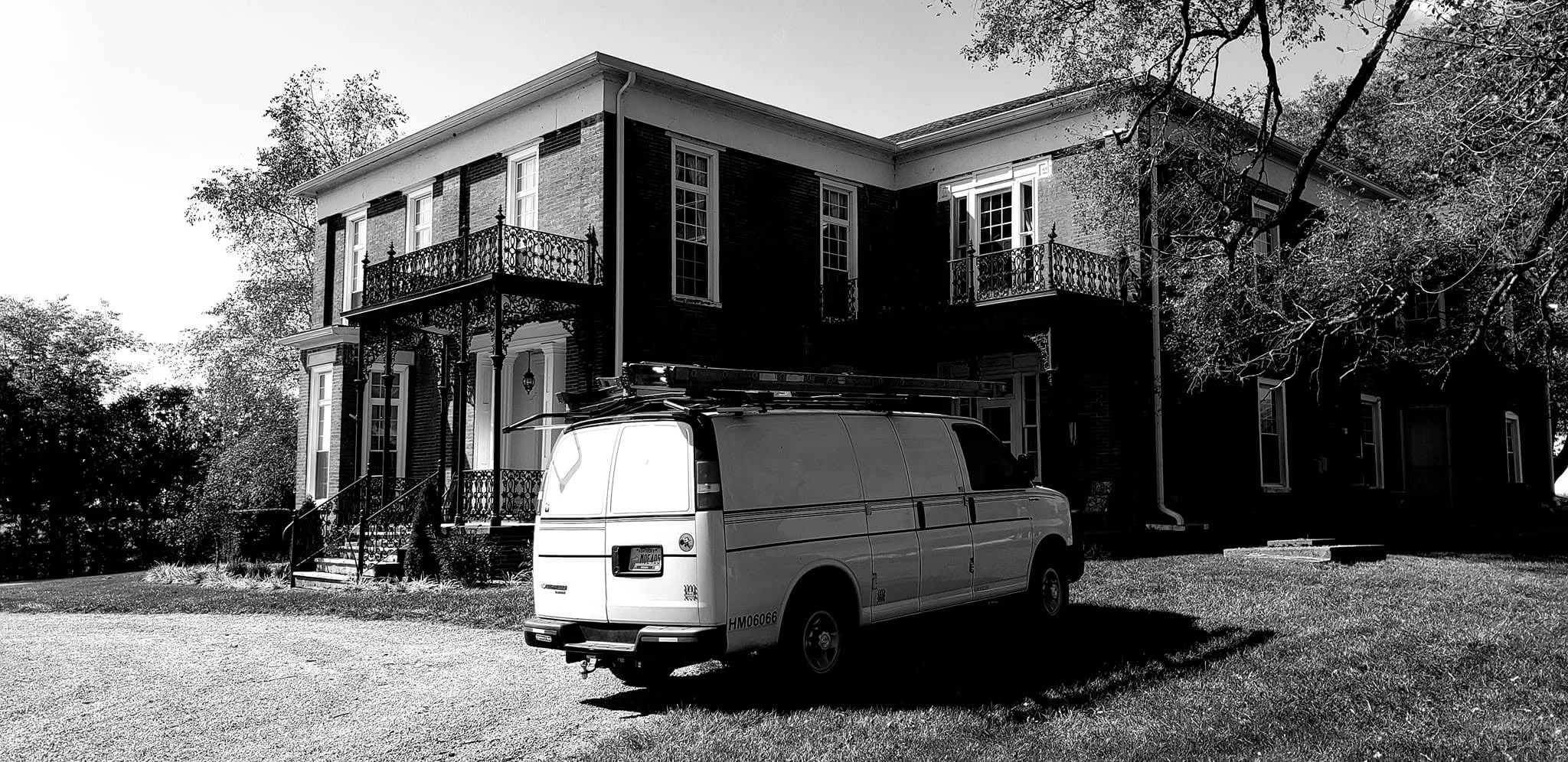 A two-story house with a brick exterior featuring a balcony and multiple windows. A white van is parked in front of the house on a grassy area, with trees surrounding the property.