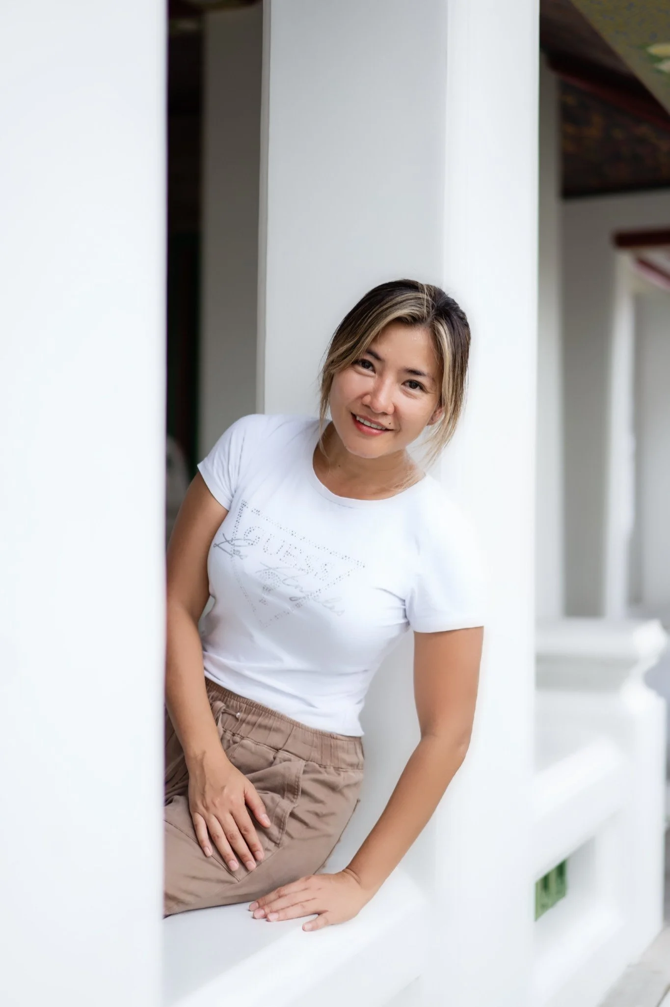 Smiling woman in casual white t-shirt and beige pants leaning on a white ledge, representing relaxation and wellness at Royal Thai Massage & Spa in Oklahoma City, serving Roberts–Crest, Belle Isle, Glenbrook, Penn Square, Classen Curve, Nichols Hills
