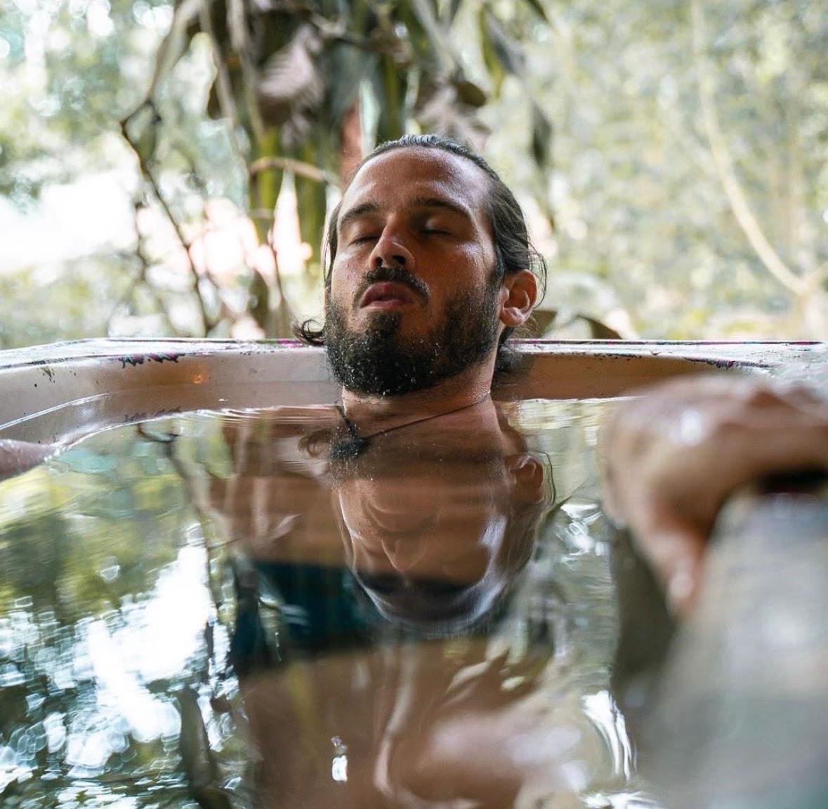 Bearded man relaxing with his eyes closed in a clear outdoor tub surrounded by trees