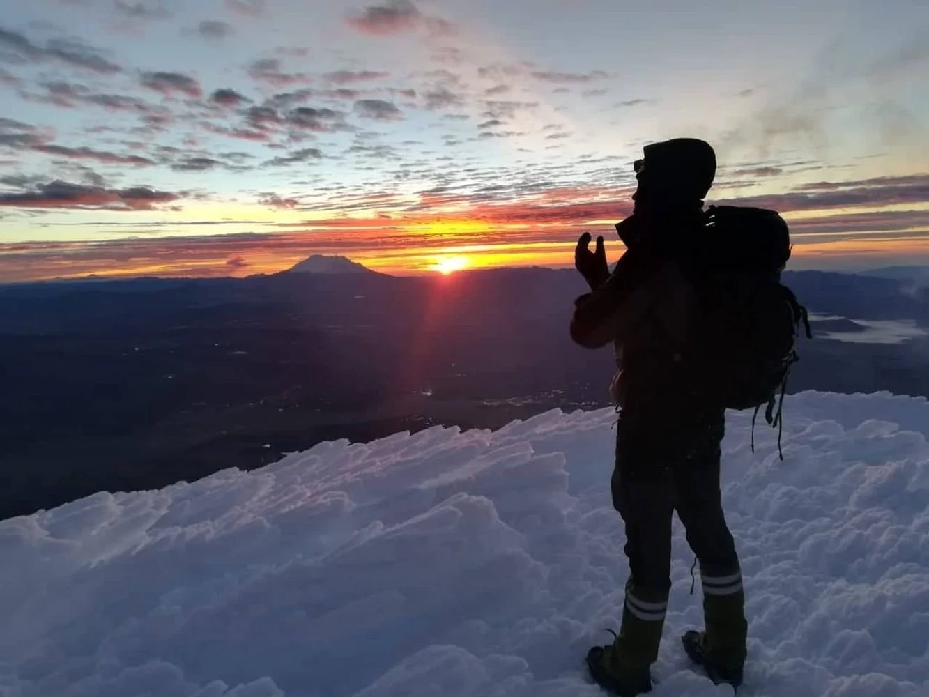 Silhouetted mountaineer with a backpack standing on snowy terrain at sunrise, overlooking distant mountains and clouds.