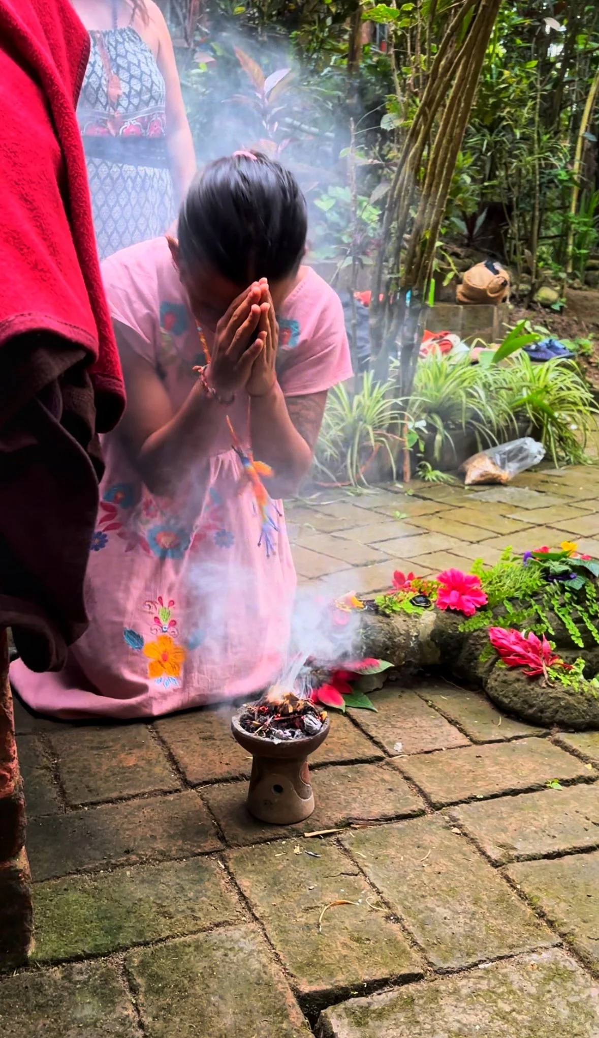 Child praying beside a smoking incense burner in a lush tropical garden with flowers
