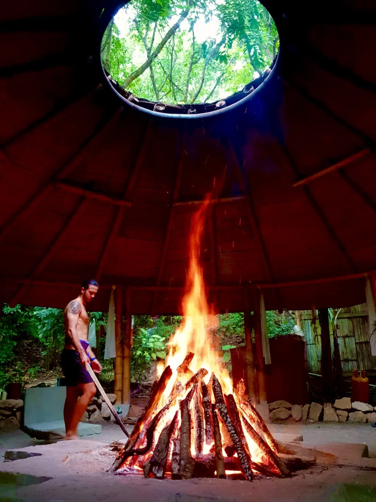 Large fire burning inside a rustic jungle shelter, with a shirtless man tending the flames and trees visible through the roof opening