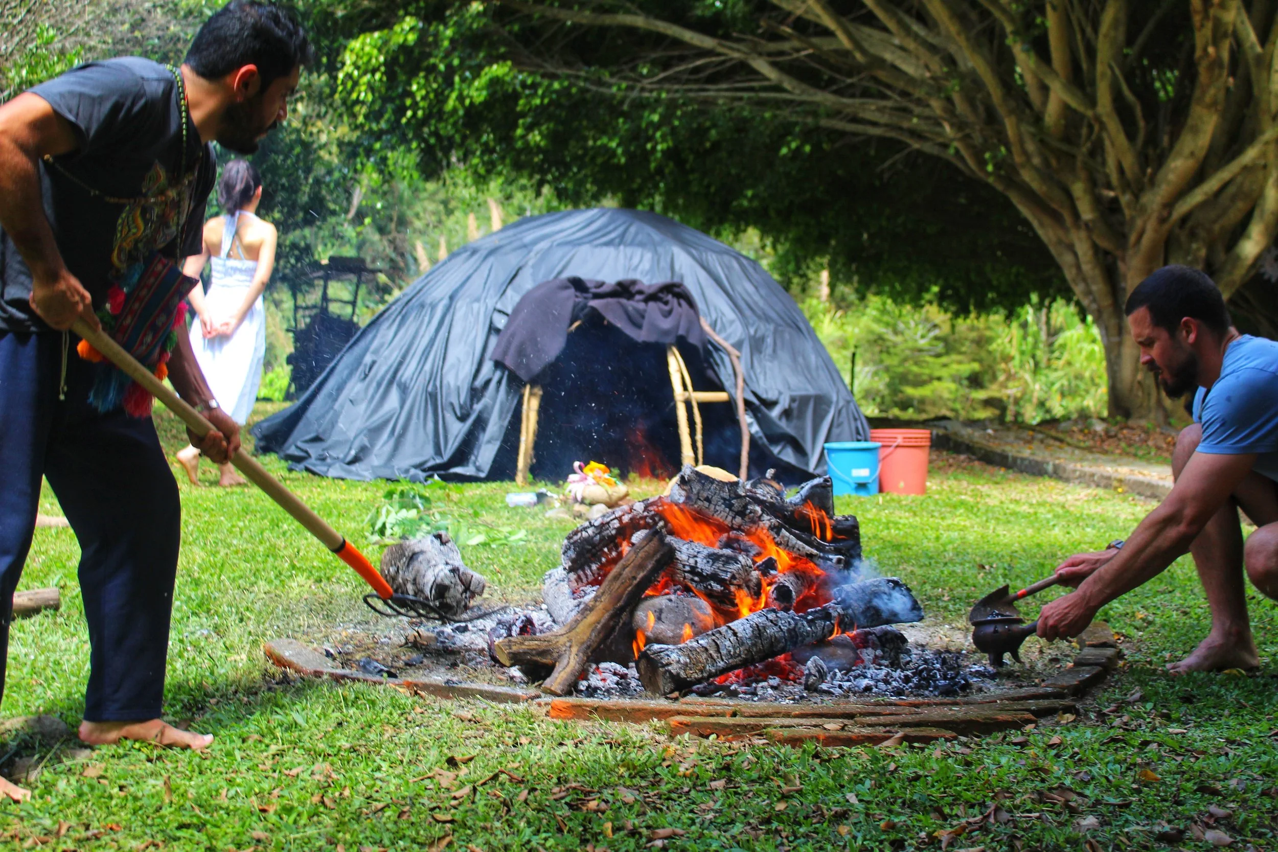 People tend a large outdoor campfire beside a black tarp shelter in a wooded area.