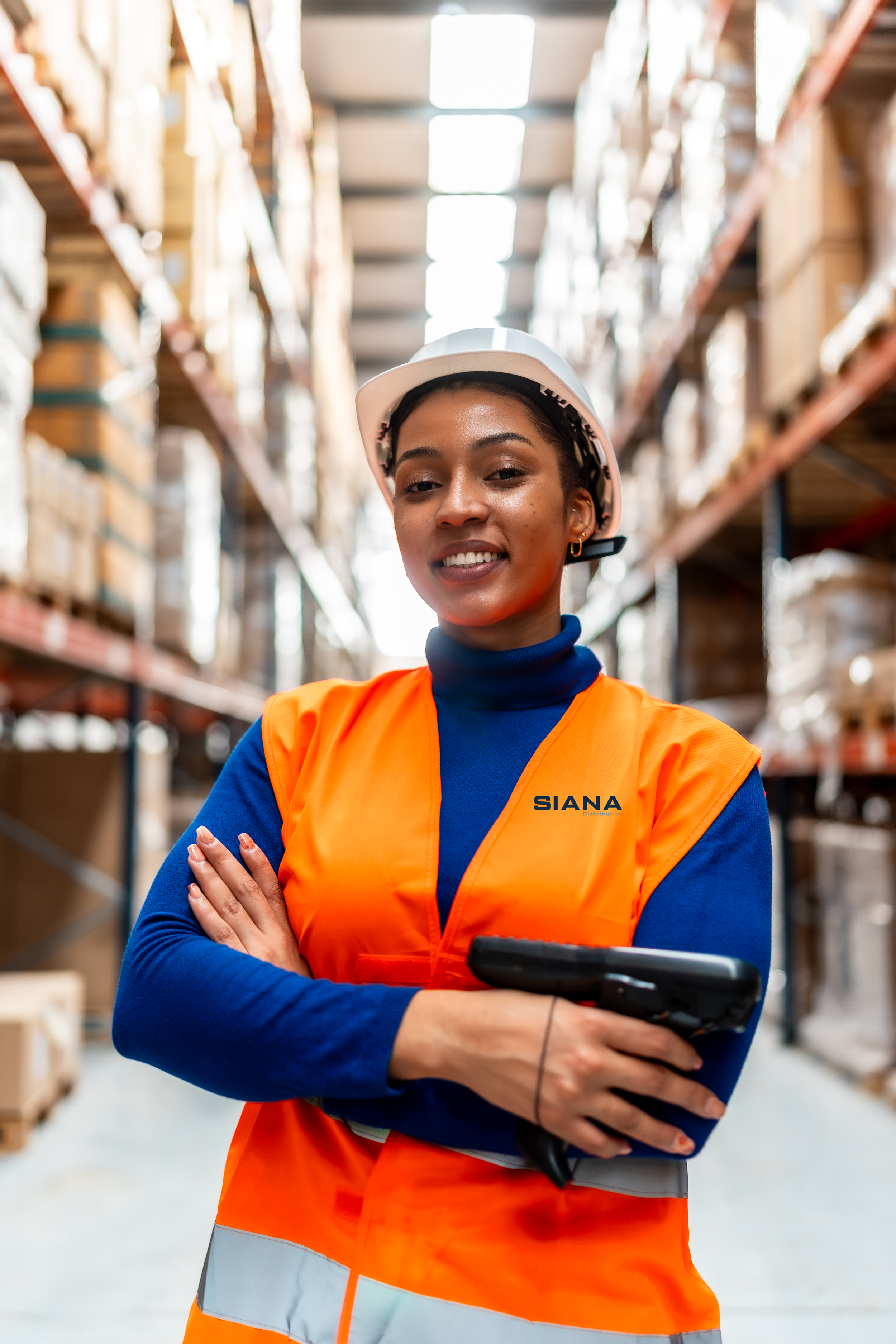 A smiling woman wearing an orange safety vest with the logo 'SIANA' and a white hard hat, standing in a warehouse aisle with shelves filled with boxes.