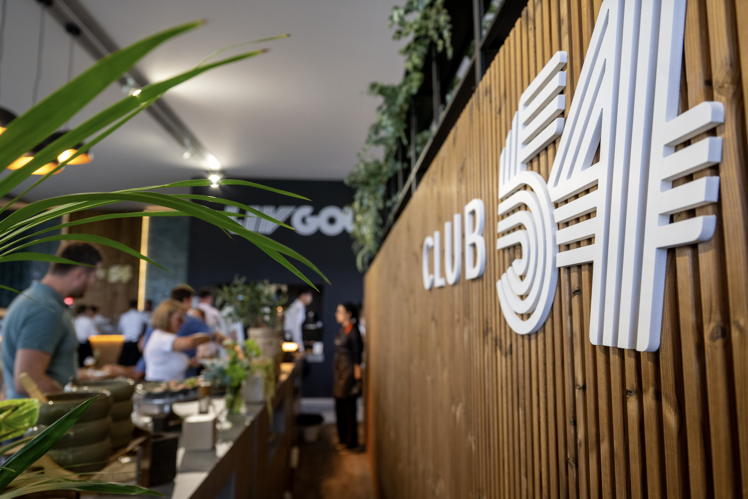 Indoor event with a wooden wall displaying a white 3D sign, greenery, and people dining in the background.