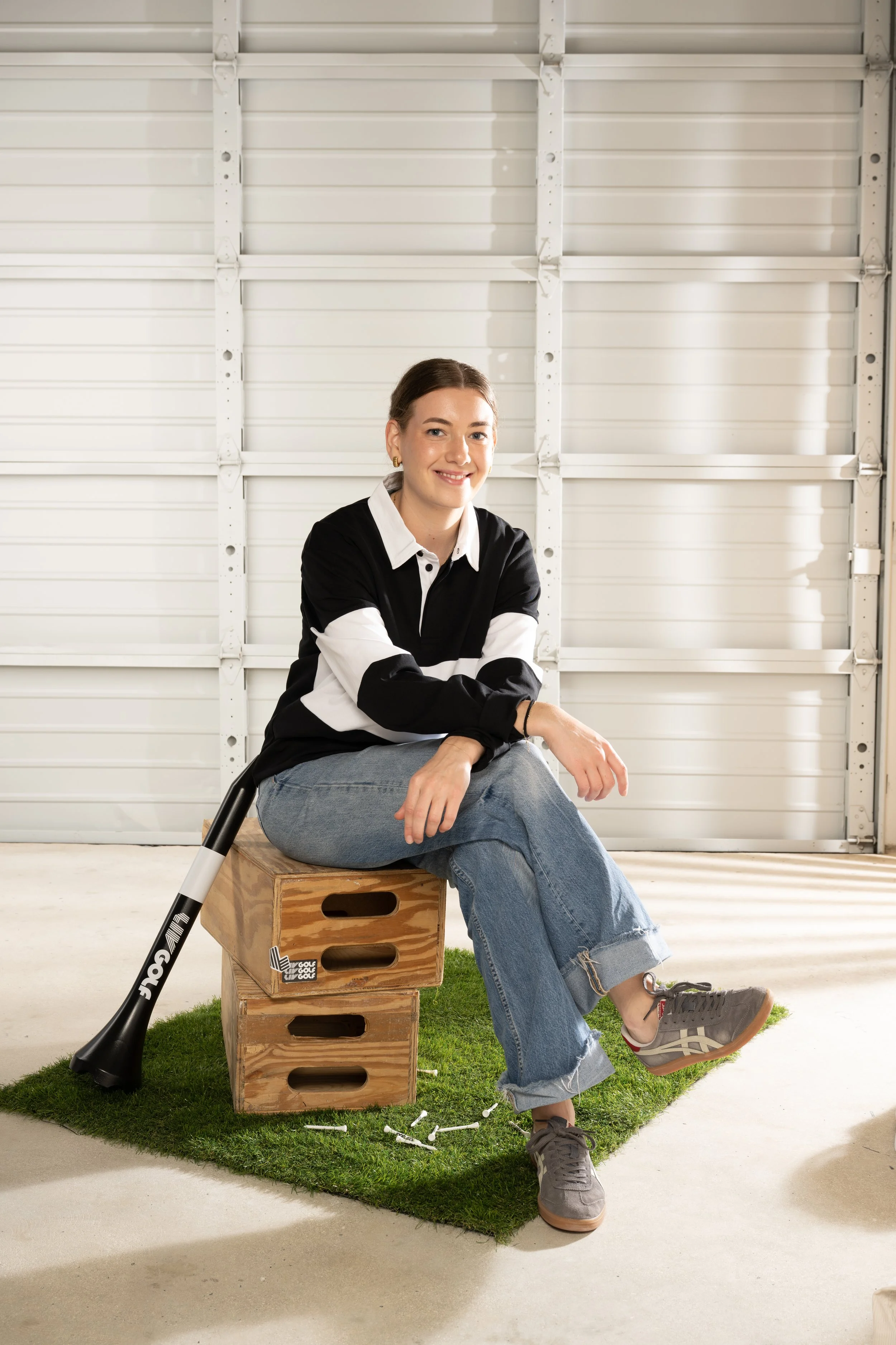 A woman sitting on wooden crates in an indoor space with a white garage door in the background. She is smiling, wearing a black and white shirt, jeans, and sneakers. There is a golf club leaning against the crates, and small white golf ball markers scattered on a small patch of artificial grass on the floor.