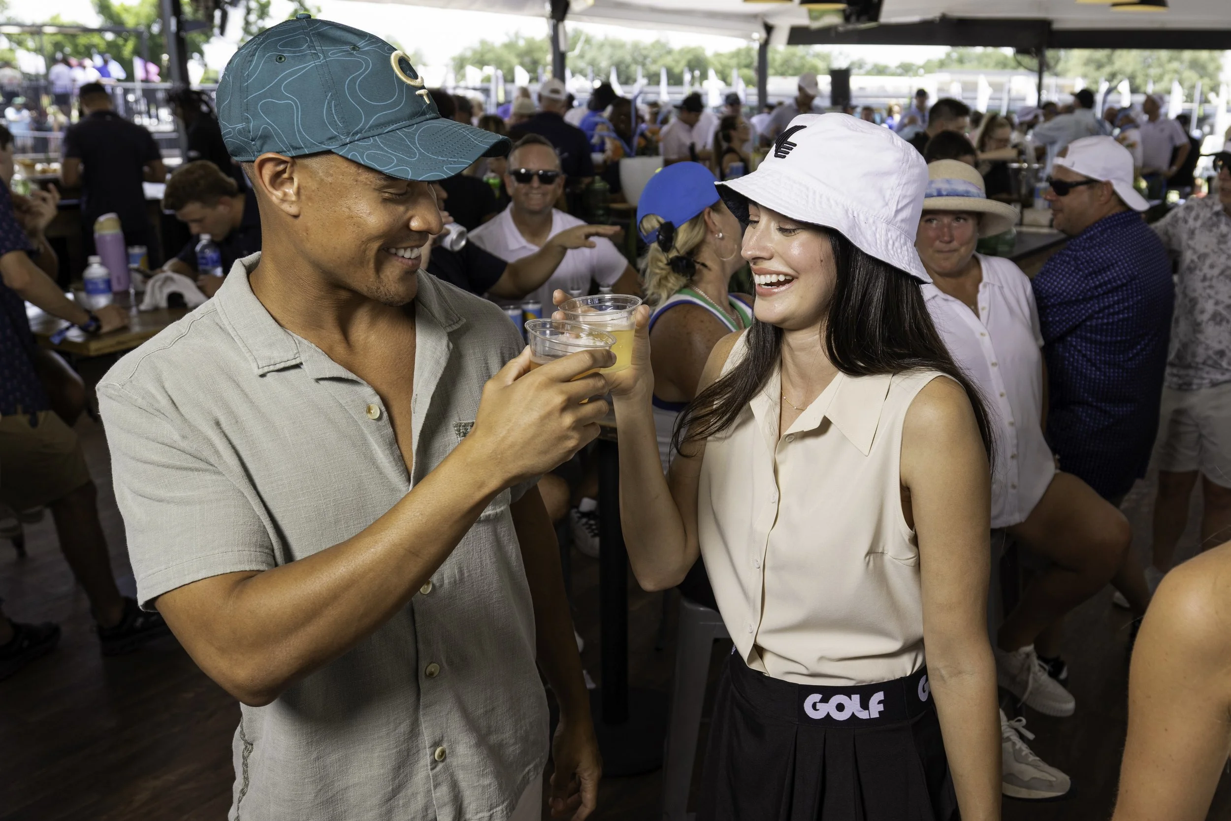 Two people, a man and a woman, smiling and clinking clear cups of beer at a crowded outdoor event under a large tent. The man is wearing a green cap and a light-colored shirt, while the woman is wearing a white bucket hat and a sleeveless top.