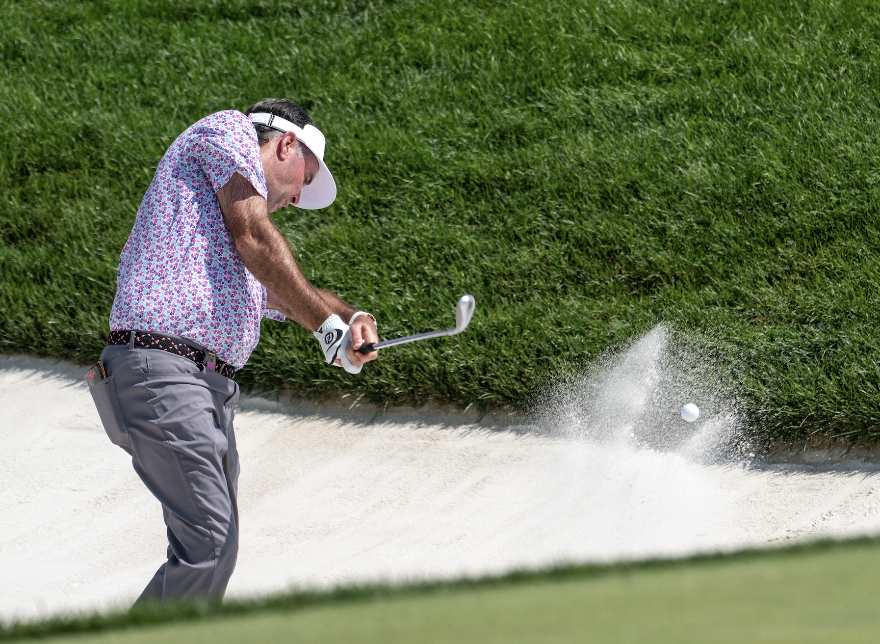 A man hitting a golf ball out of a sand bunker on a golf course, with sand flying in the air, green grass in the background, and wearing a floral shirt, gray pants, and a white cap.
