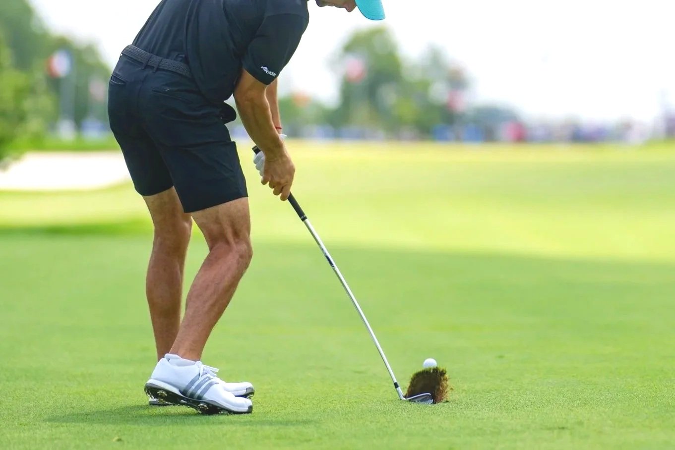 A man wearing a black shirt, black shorts, white shoes, and a blue cap preparing to tee off on a golf course with a golf club, a golf ball, and a tee.