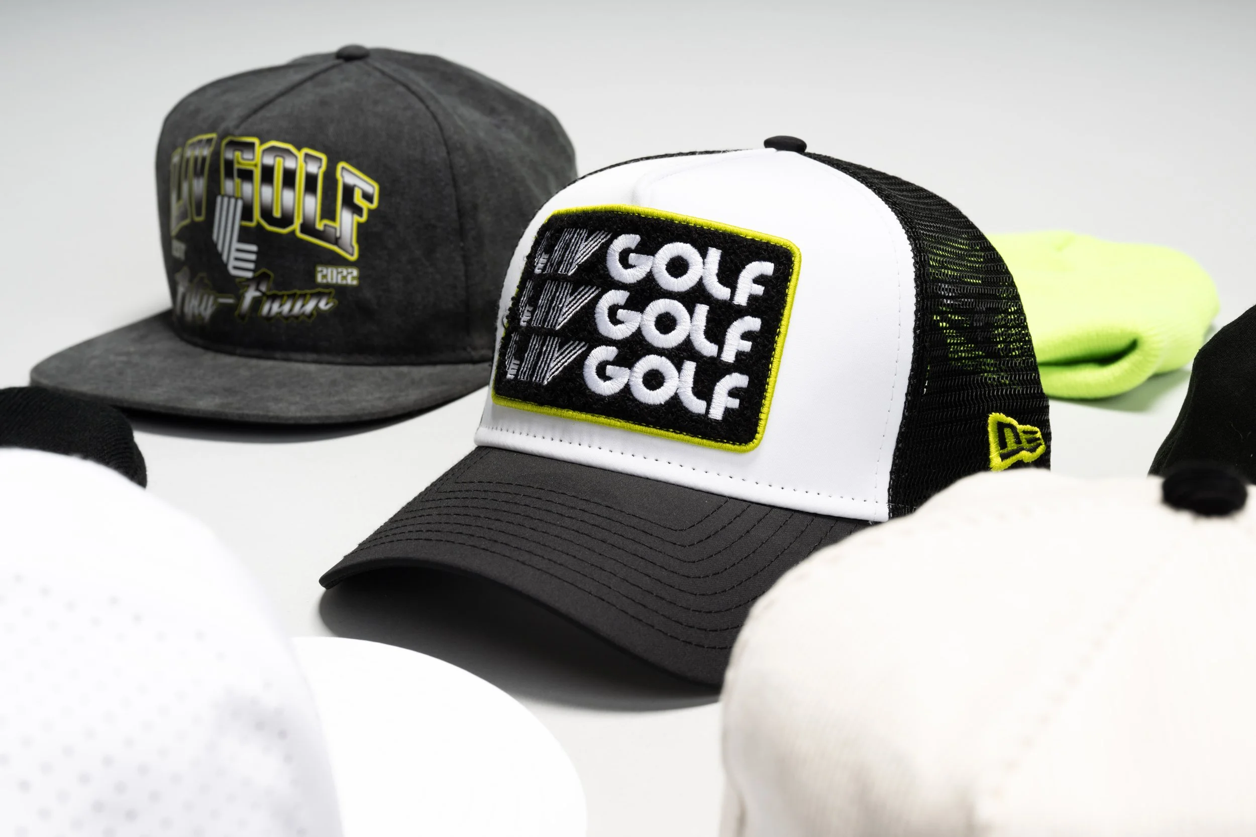 Close-up of two golf caps, one gray and one black and white, with patches that say "GOLF" and include a barcode design. There is also a neon yellow-green object in the background, possibly a glove, and some white fabric or additional clothing items nearby.