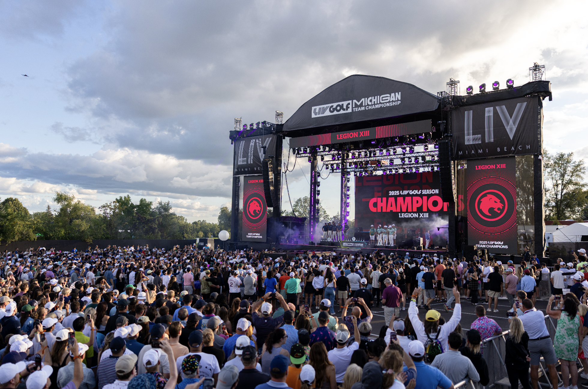 A large crowd of people gathered outdoors at a golf event with a stage in the background. The stage displays banners for the 2025 golf season champion, Legion XIII, with branding for the Michigan Team Championship and Miami Beach. The sky is partly cloudy, and the crowd is wearing casual clothing and hats.
