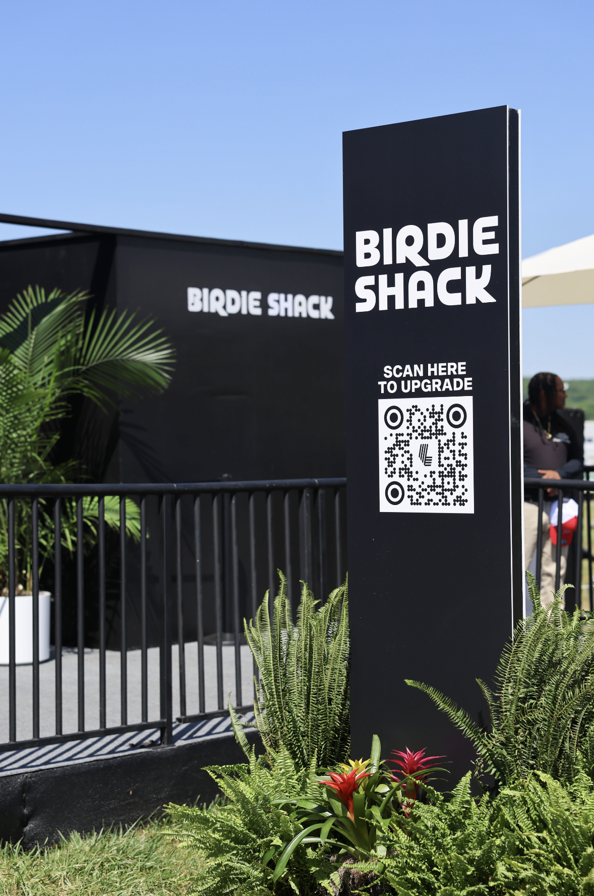 Sign for Birdie Shack with QR code, outdoor setting with plants in foreground, black fence, black tent in background, and a person sitting under an umbrella.