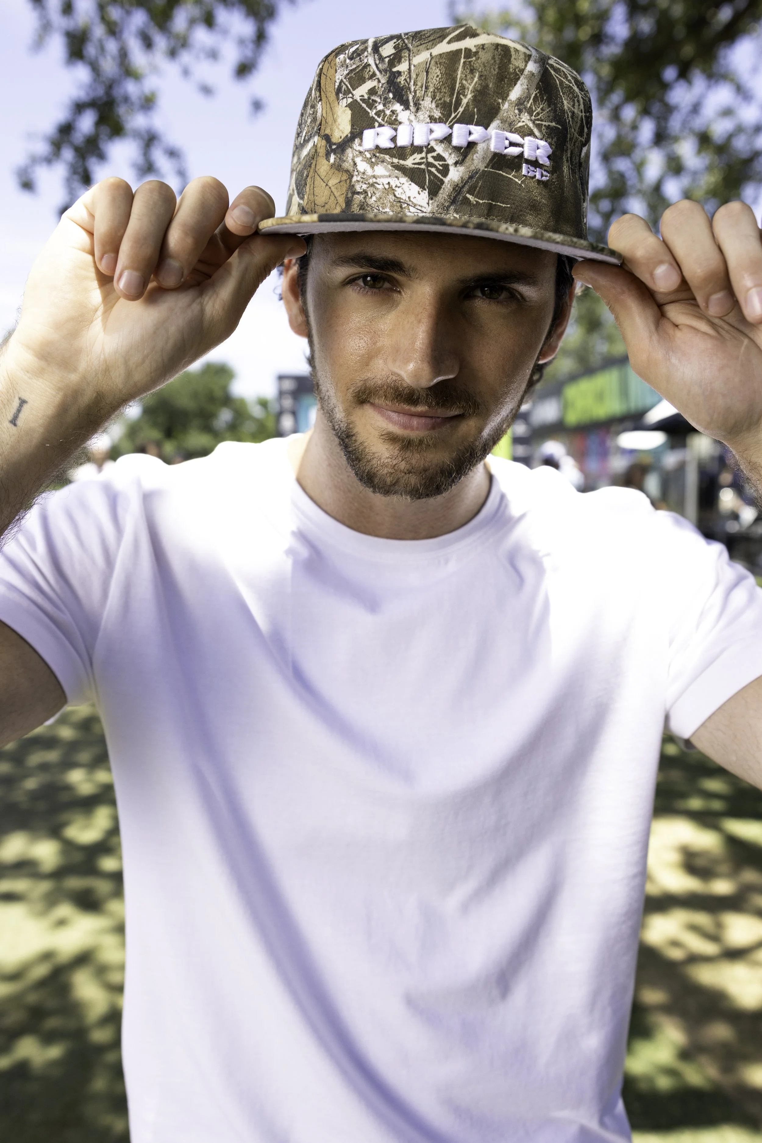 A young man with a beard, wearing a white t-shirt and a camouflage baseball cap with 'RIDER' embroidered on it, standing outdoors in a park with trees and a blue sky.