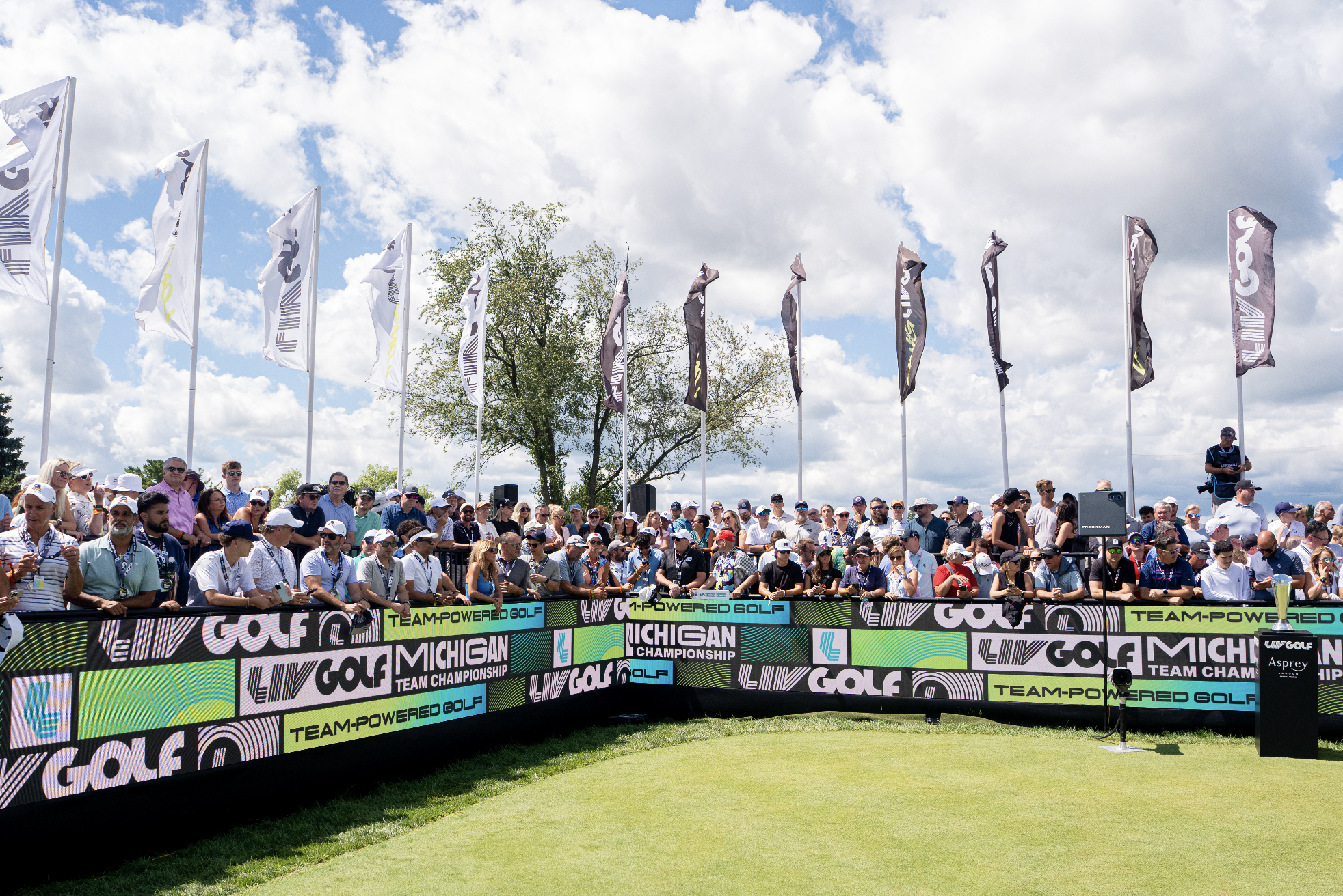 Crowd of spectators watching a golf tournament outdoors on a sunny day under a partly cloudy sky, with flags and banners displaying "GOLF," "TEAM-POWERED GOLF," and "MICHIGAN."