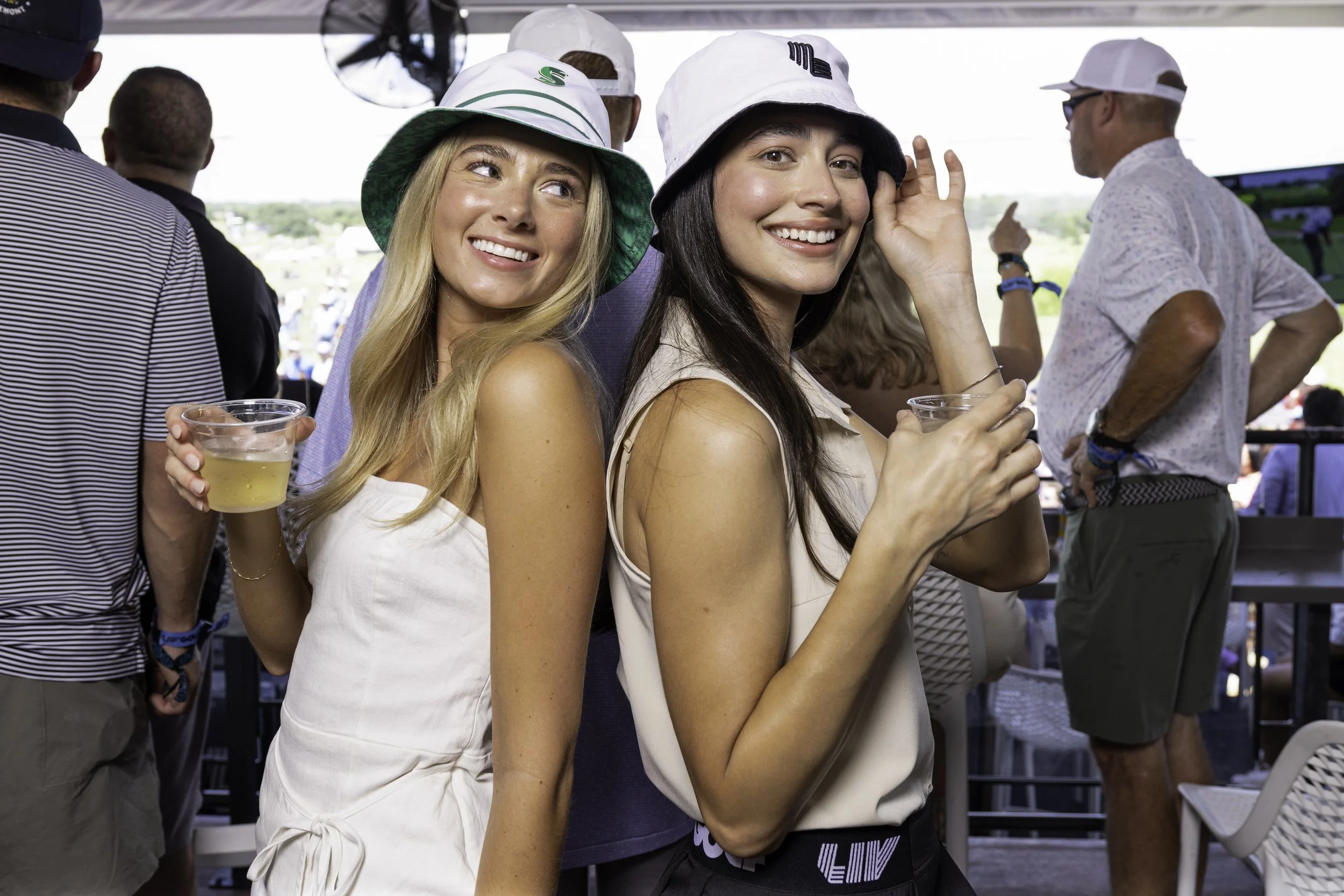 Two young women at an outdoor social event, smiling and holding drinks, wearing casual summer attire and hats.